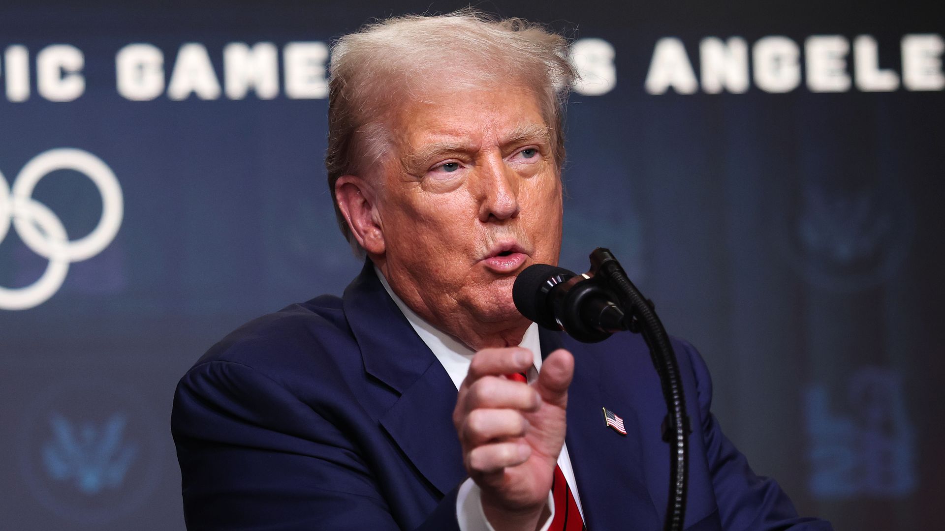 President Trump, wearing a dark suit, white shirt, and red tie speaks into a microphone against a backdrop featuring Olympic rings and the words "LOS ANGELES".