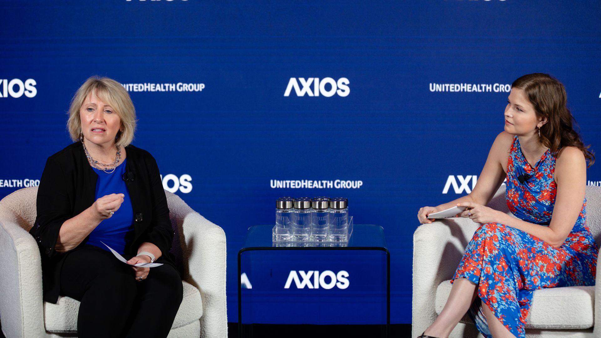 Two women sit in white chairs on a panel stage against a blue backdrop with Axios and UnitedHealth Group logos; a glass table with water bottles sits between them.