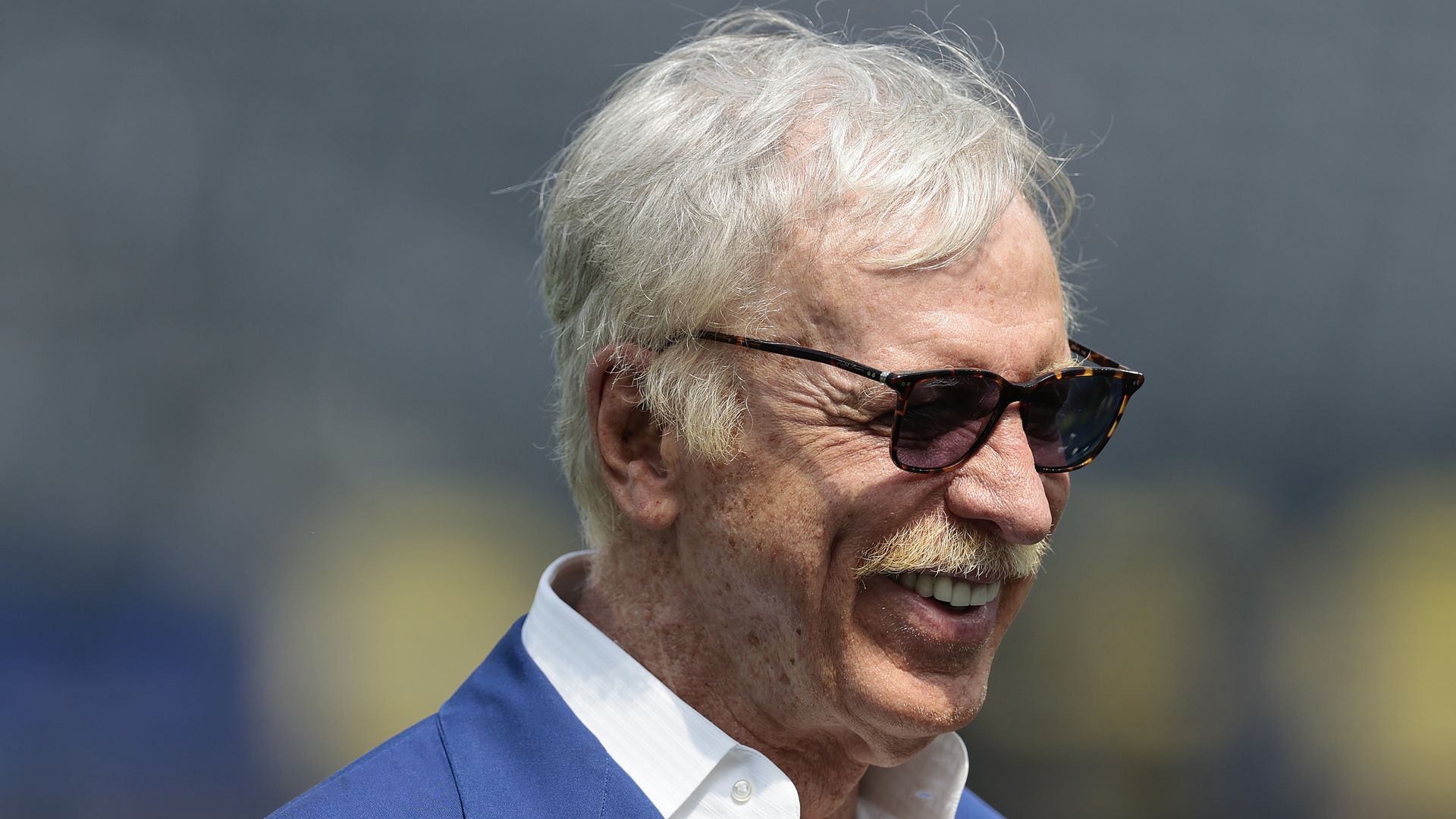Close-up of a man with white hair and a mustache wearing tortoiseshell sunglasses and a blue suit jacket over a white shirt, smiling outdoors.