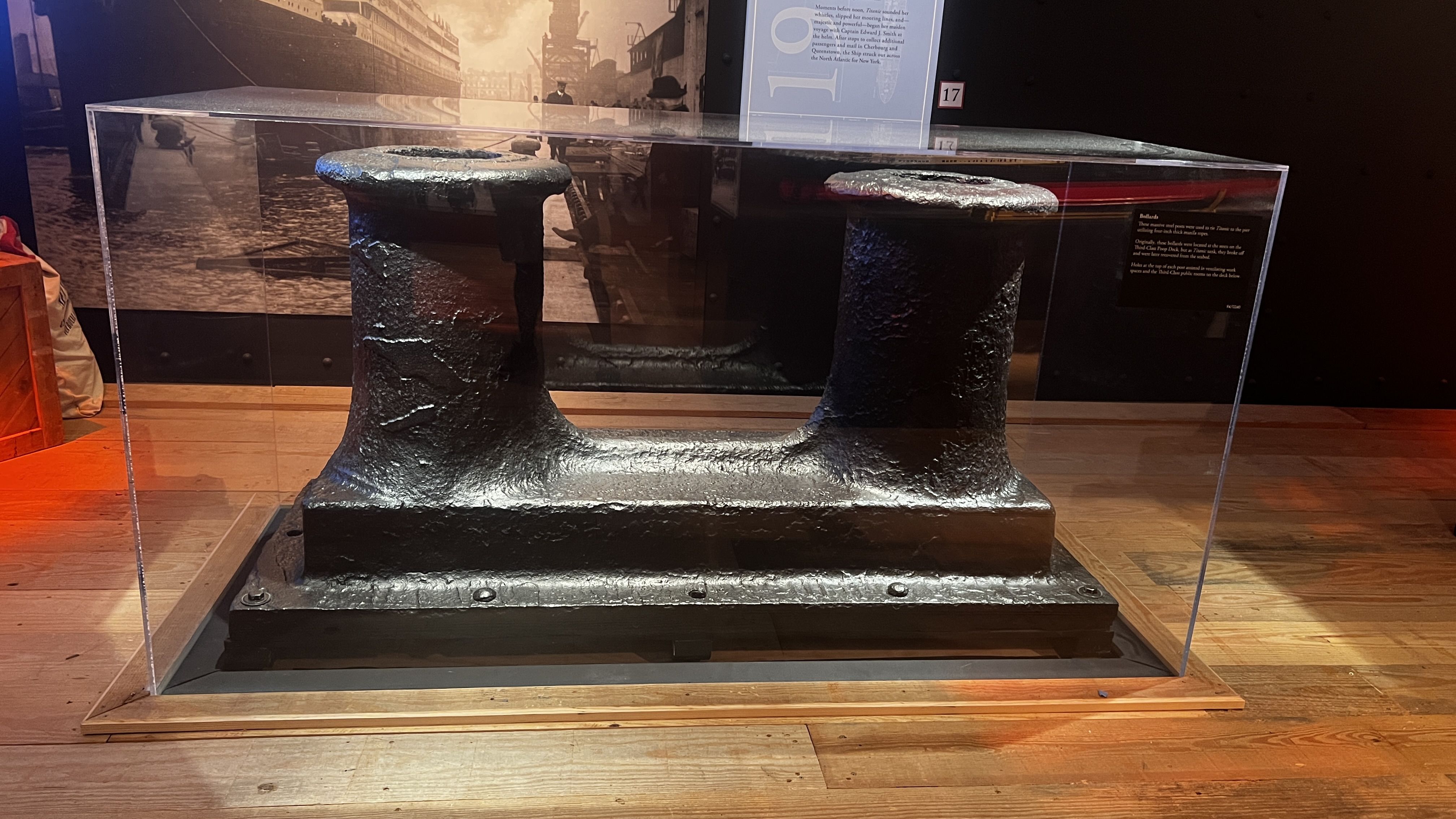 Large black iron artifact inside a glass display case. Two thick vertical columns with circular tops are linked by a curved piece, set on a wooden museum floor.