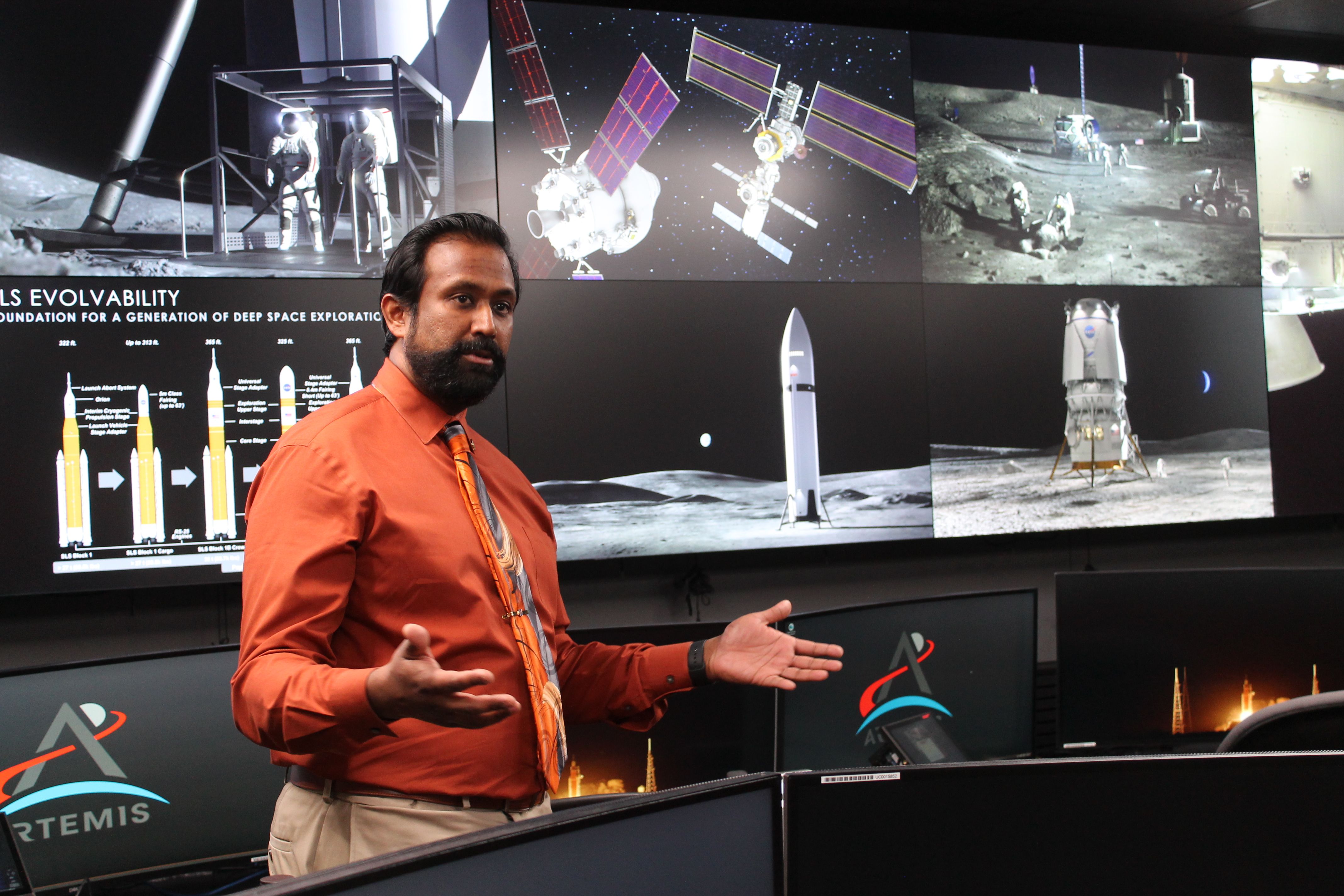 Man in orange shirt and orange space-themed tie giving presentation in front of screens showing lunar and space exploration images, including astronauts, rockets, and satellites, labeled Artemis.