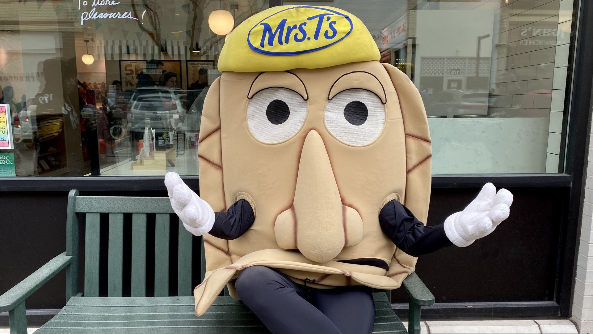 Mascot in a beige nose costume sits on a green bench outside a shop, wearing a yellow cap with a blue "Mrs. T's" logo, white gloves, black sleeves, and yellow sneakers; arms open.