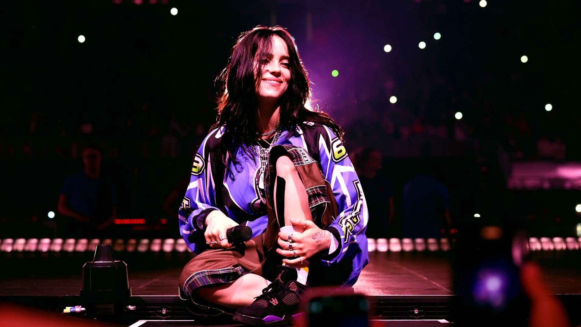 A performer with long dark hair smiles while sitting cross-legged on stage during a concert, holding a microphone. She wears a blue oversized jersey and brown plaid shorts, with the audience and stage lights glowing behind her.