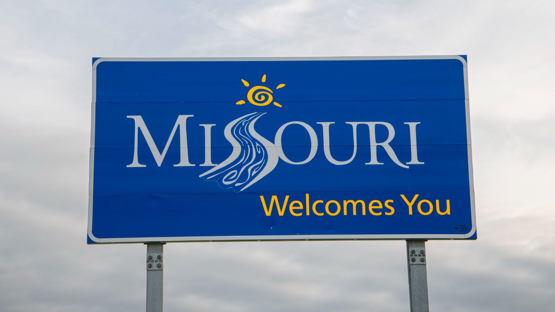 A blue sign with the words "Missouri Welcomes You" in white and yellow against an overcast sky.