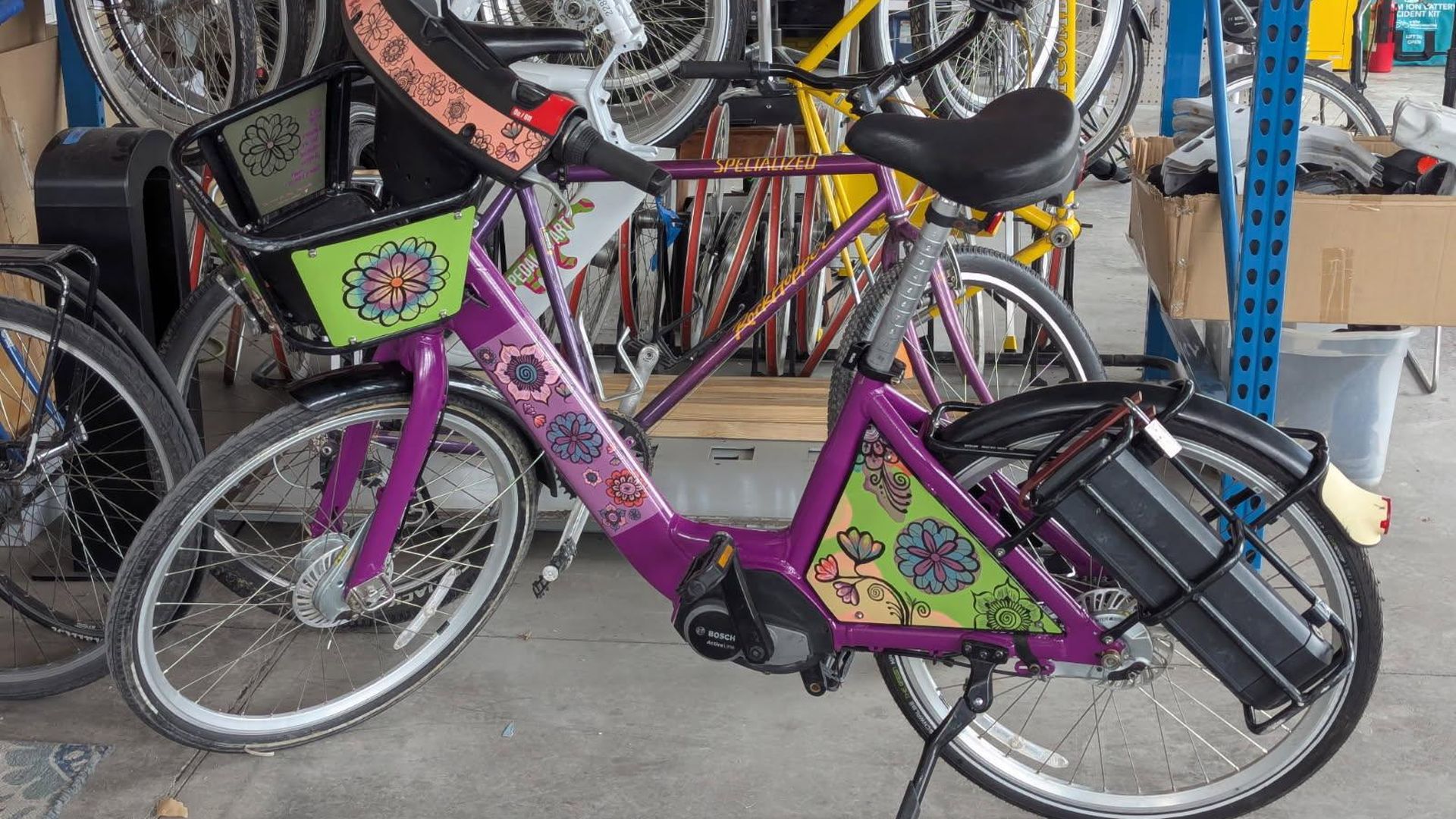 A purple bicycle with colorful flower decals and green panels, parked in a busy bike shop among racks and other bikes.