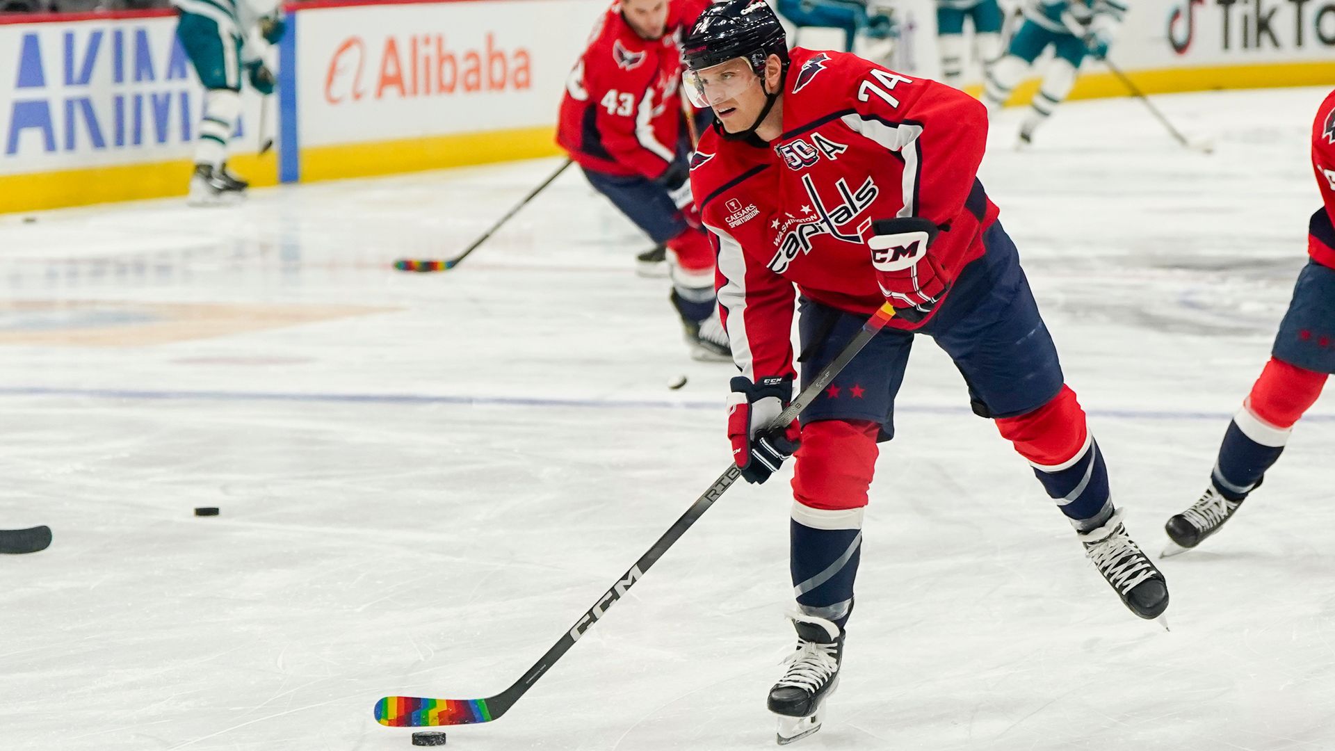 Hockey player in red Washington Capitals jersey with number 74 skating on ice, holding stick with rainbow tape, focus on puck. Other players and advertisements visible in background.