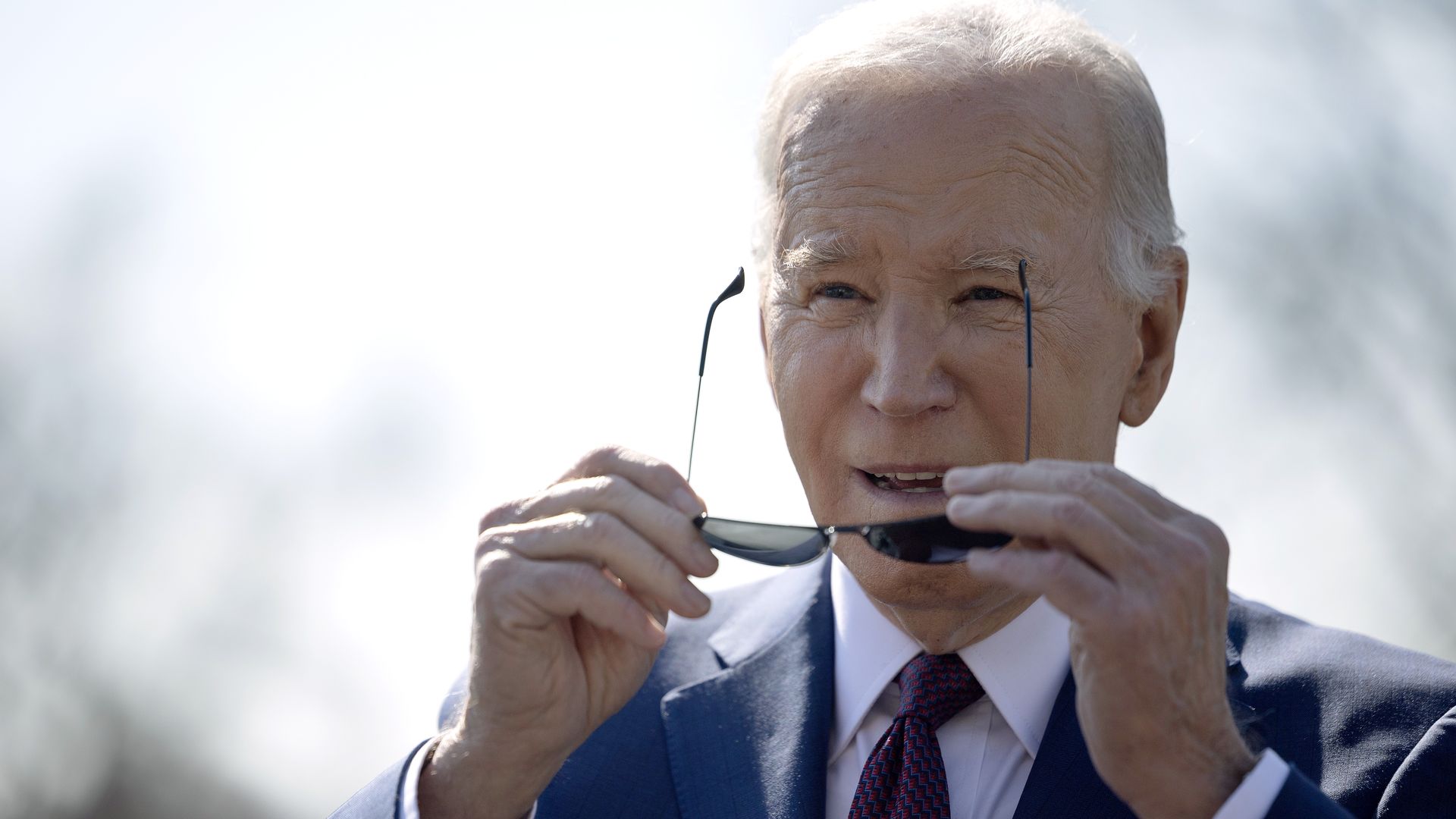 President Joe Biden, wearing a blue suit, white shirt and red and blue tie, puts on aviator sunglasses while outside.