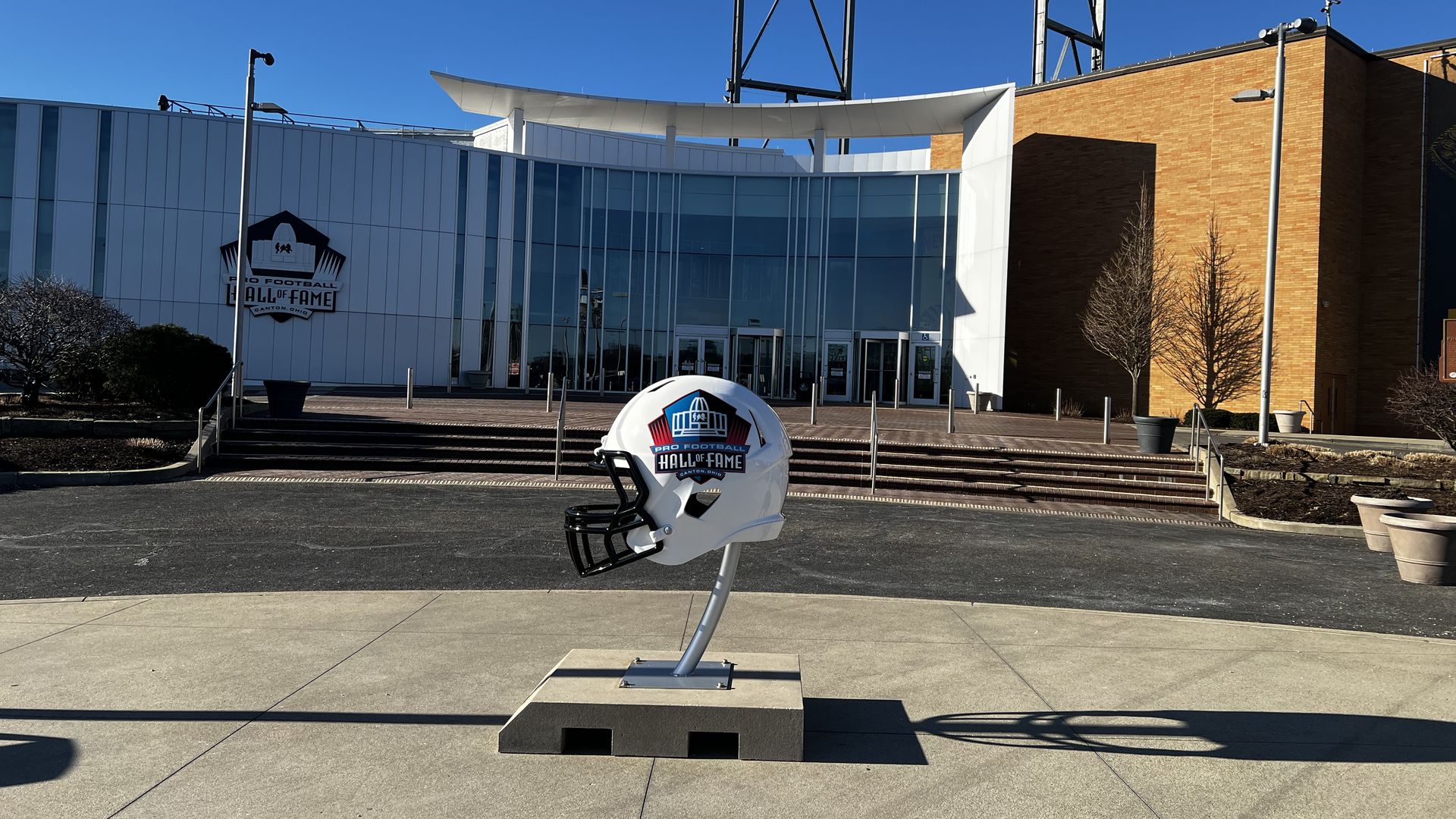 White football helmet sculpture with "Pro Football Hall of Fame" logo in front of a modern building with glass entrance and a blue sky.