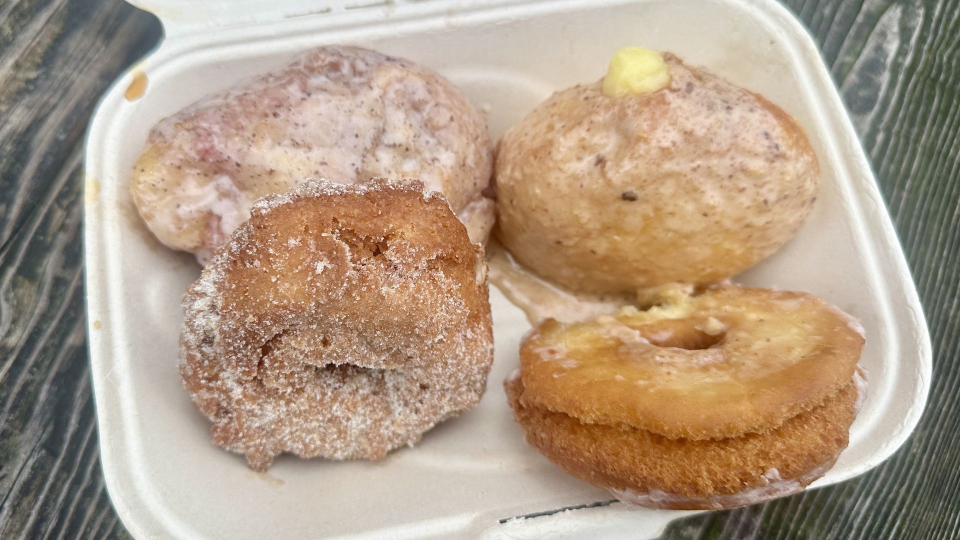 Four doughnuts in a to-go container, including a fritter, two cake doughnuts and a filled doughnut with lemon curd peeking out..