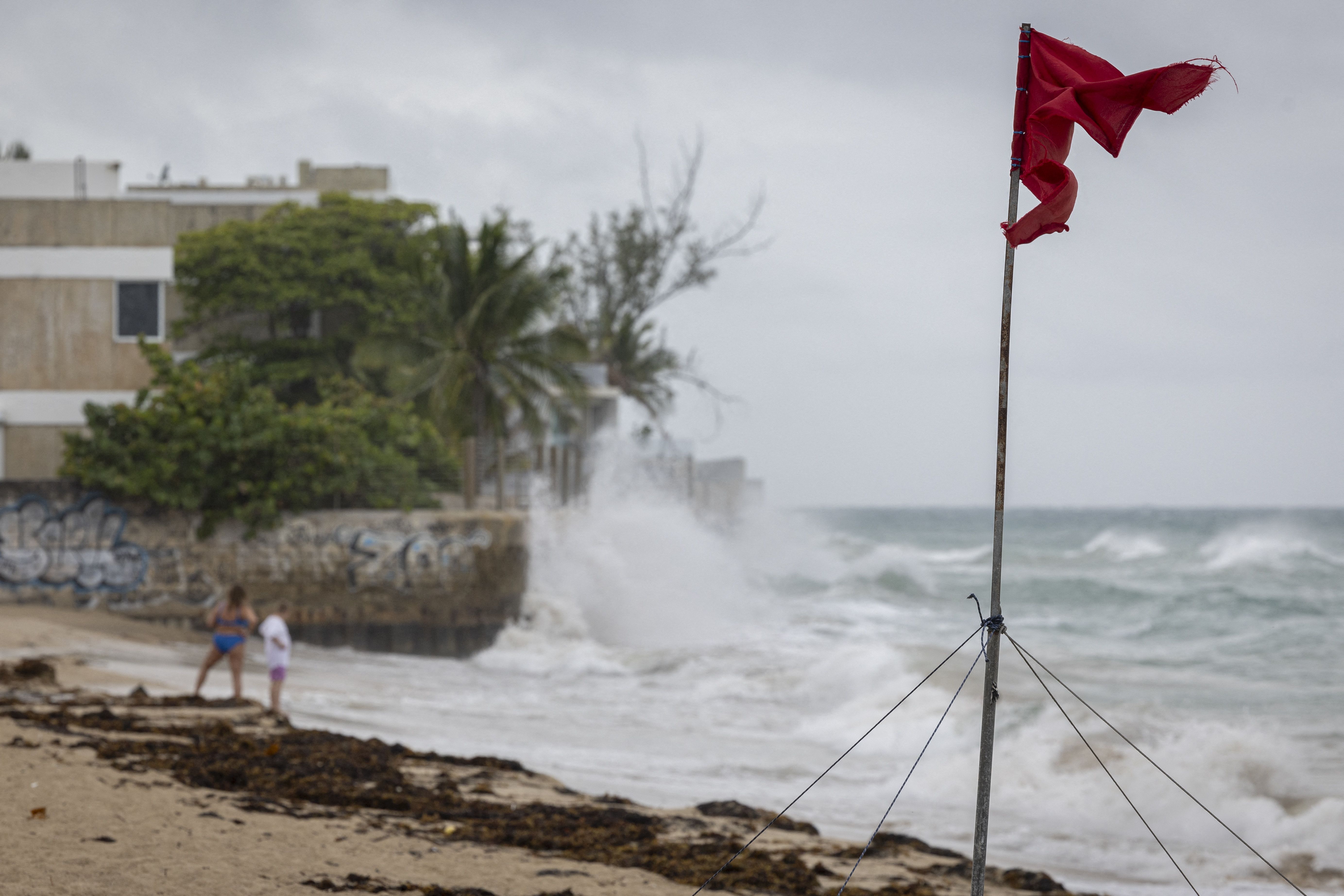Torn red warning flag on beach near rough ocean waves crashing against a seawall with two people standing on sand, gray sky, and greenery in background.
