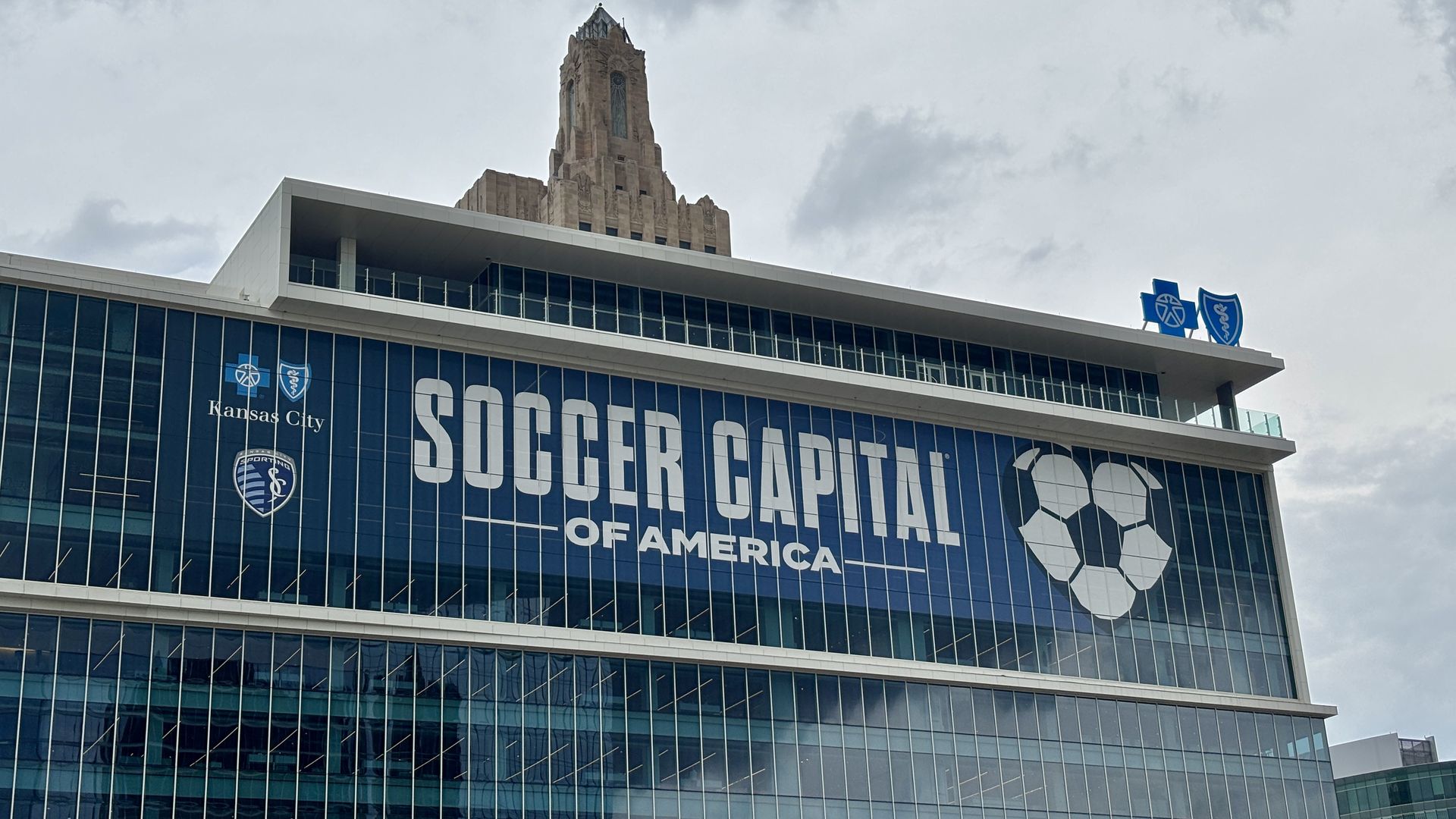 Modern glass stadium facade with a large blue banner reading "SOCCER CAPITAL OF AMERICA" and Kansas City logos; a beige Art Deco tower rises behind under a cloudy sky.