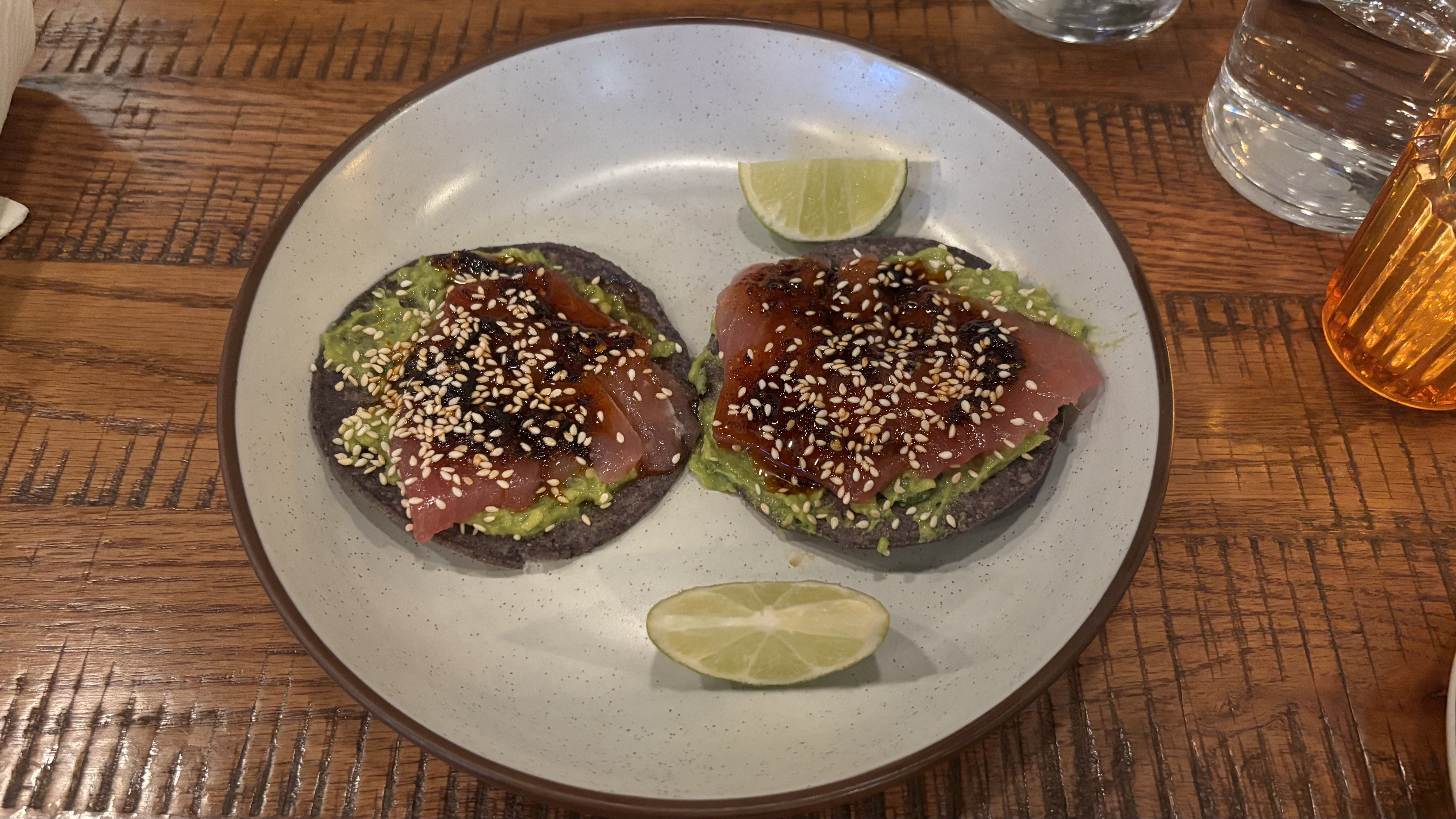Two blue corn tostadas topped with avocado and slices of seared tuna, sprinkled with sesame seeds and glazed, on a speckled plate with lime wedges on a wooden table.