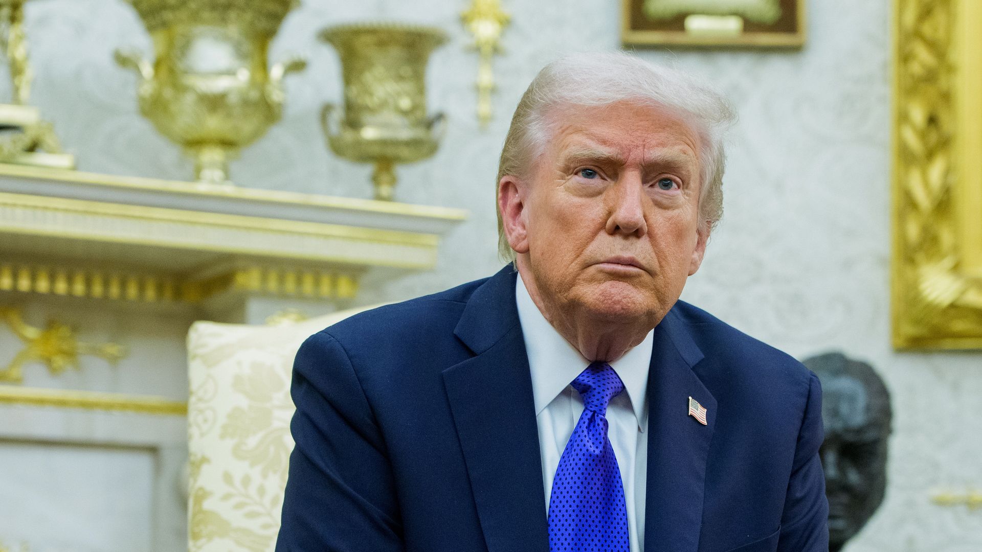 Older man with light hair in a navy suit and blue tie sitting in an ornate room with gold decorations and patterned wallpaper, looking serious.