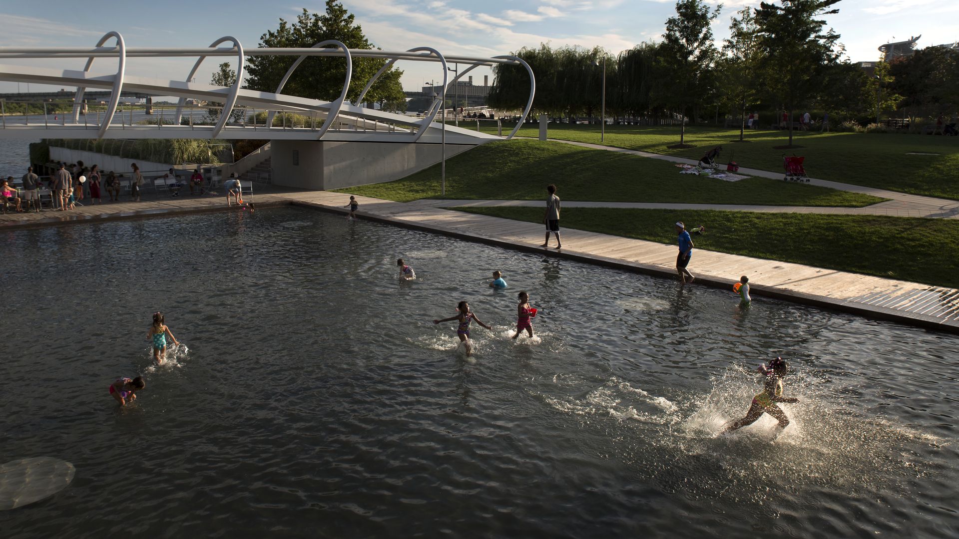 Sunlit riverside park with a futuristic white arched bridge over a shallow pool; people lounging by the water, kids swimming, grassy lawn, trees, and a skyline in the background.