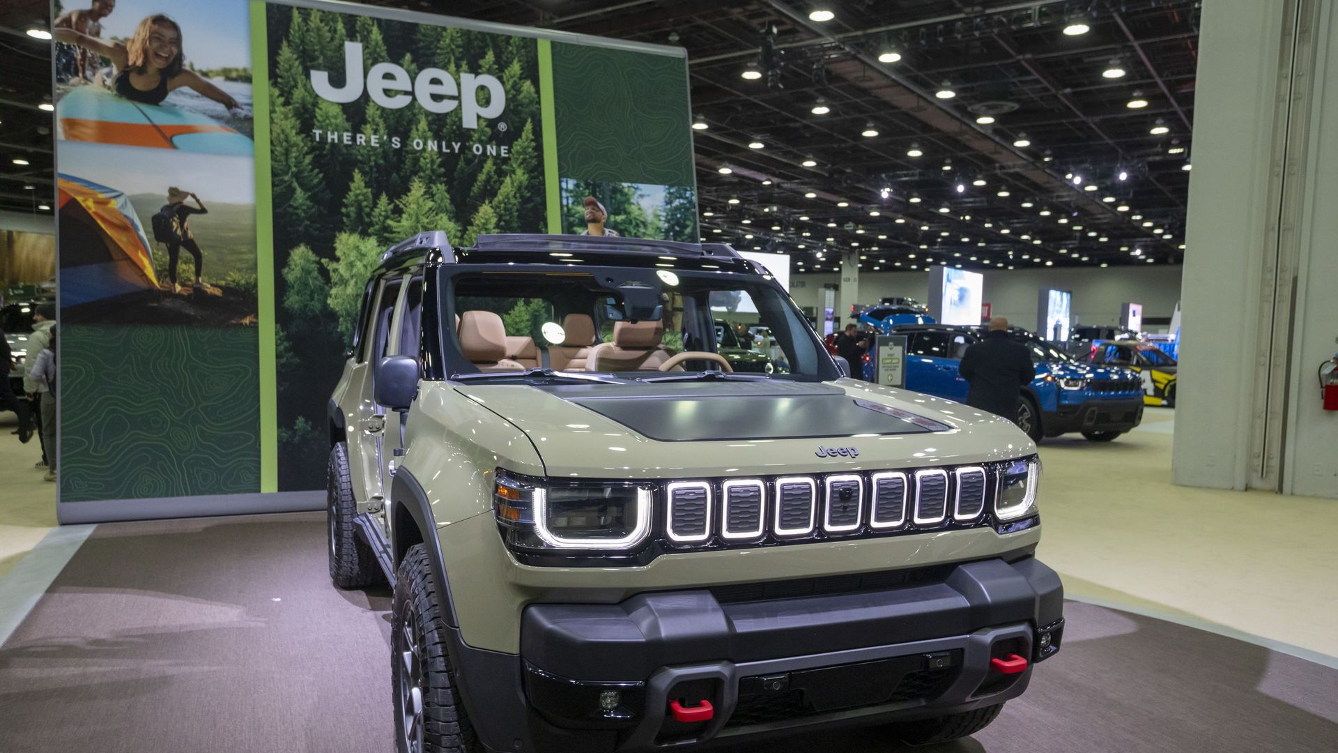 Indoor auto show display featuring a gray Jeep SUV with beige interior and red tow hooks, in front of a large Jeep banner showing a forest scene and the slogan There’s Only One.