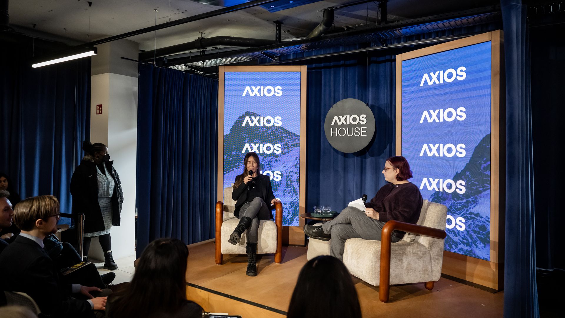 Two women speaking on a small stage at an Axios House event, with blue curtains and large screens displaying the Axios logo behind them, and an audience watching attentively.