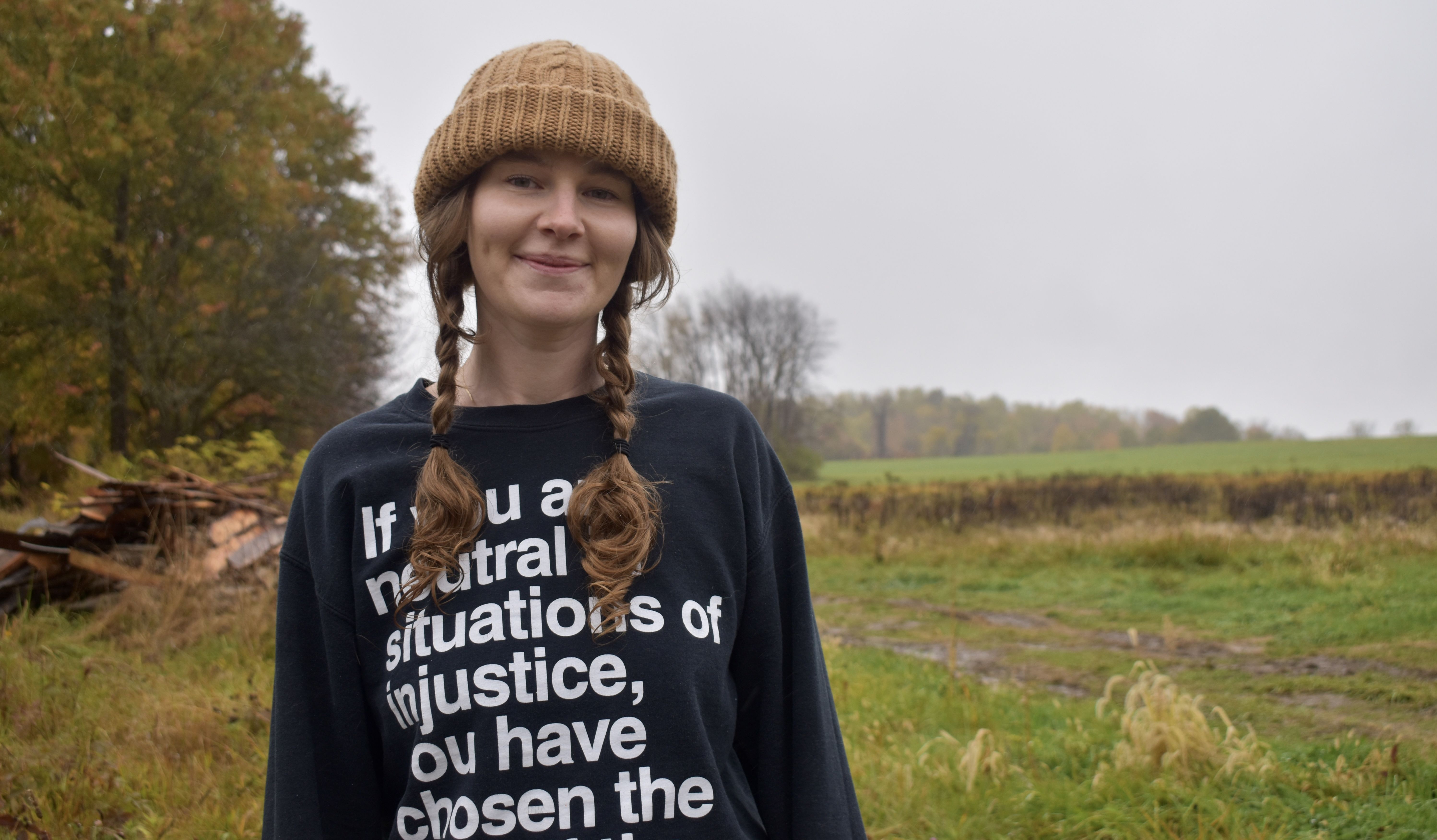 Smiling woman with two braids wearing a brown knit hat and a black sweatshirt with white text, standing outdoors on a cloudy day with autumn trees and green fields in the background.