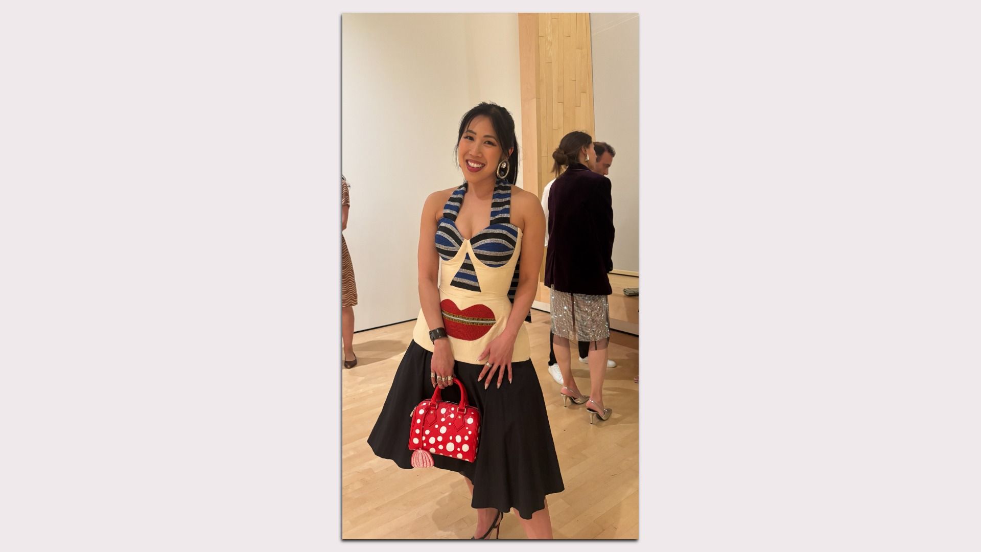Smiling woman in a colorful graphic dress with a blue striped bodice and large red lips, carrying a red polka-dotted handbag; gallery visitors in the background.