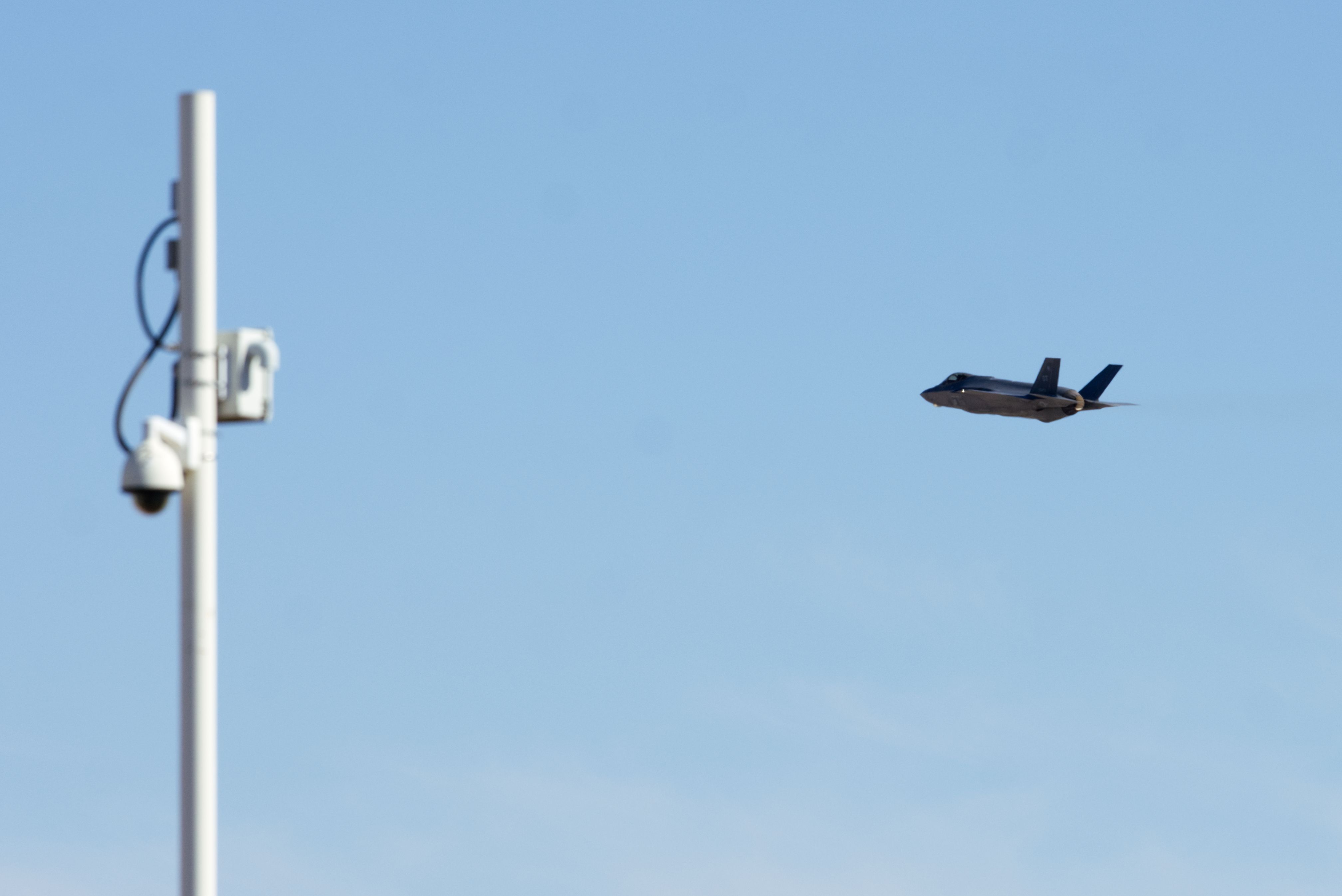 Dark gray military jet flying in clear blue sky with a white surveillance camera on a pole in the foreground to the left.