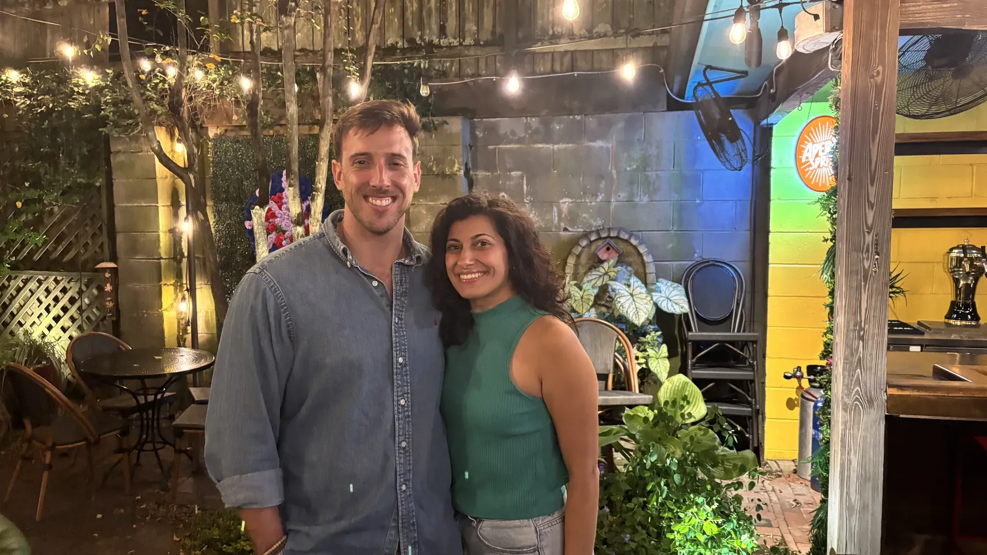 Two people smile for a photo in a cozy outdoor patio with string lights, plants, and a colorful wall. A man in a denim shirt stands beside a woman in a green sleeveless top.