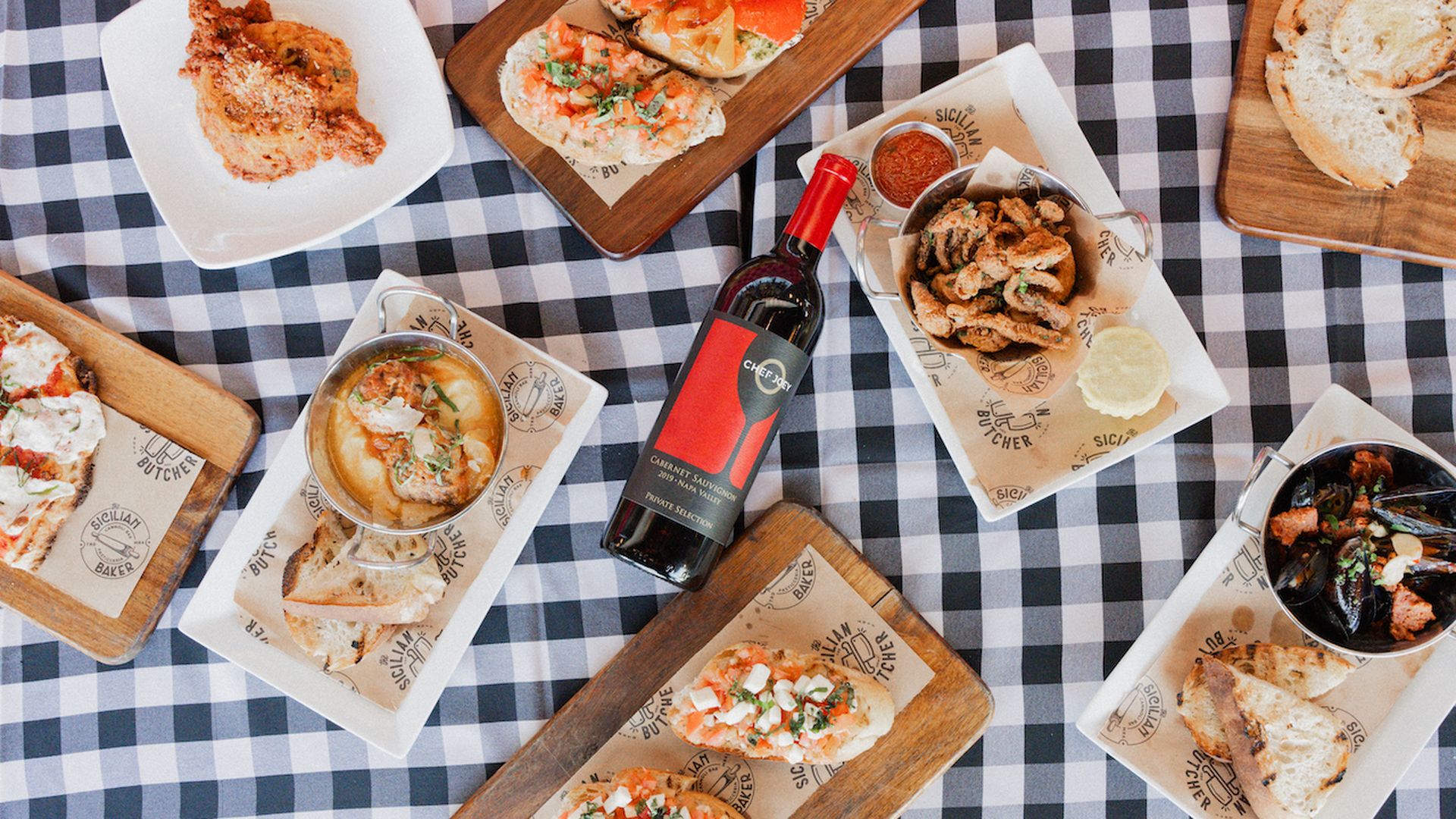 An assortment of appetizers arranged on black gingham blanket with a bottle of wine in the middle.