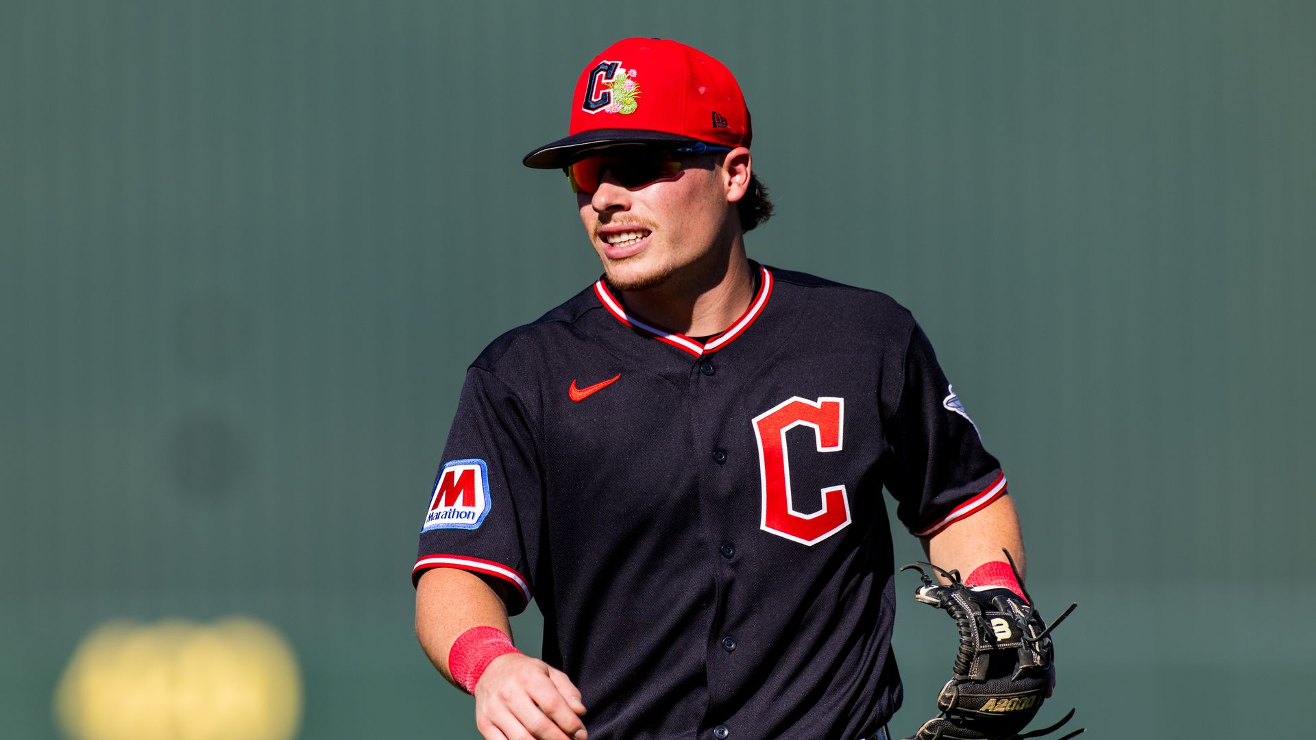 A baseball player wearing a red cap and black jersey with red trim and a large red letter C on the chest, sunglasses on, and a baseball glove in hand, standing on a field with a green outfield wall behind.