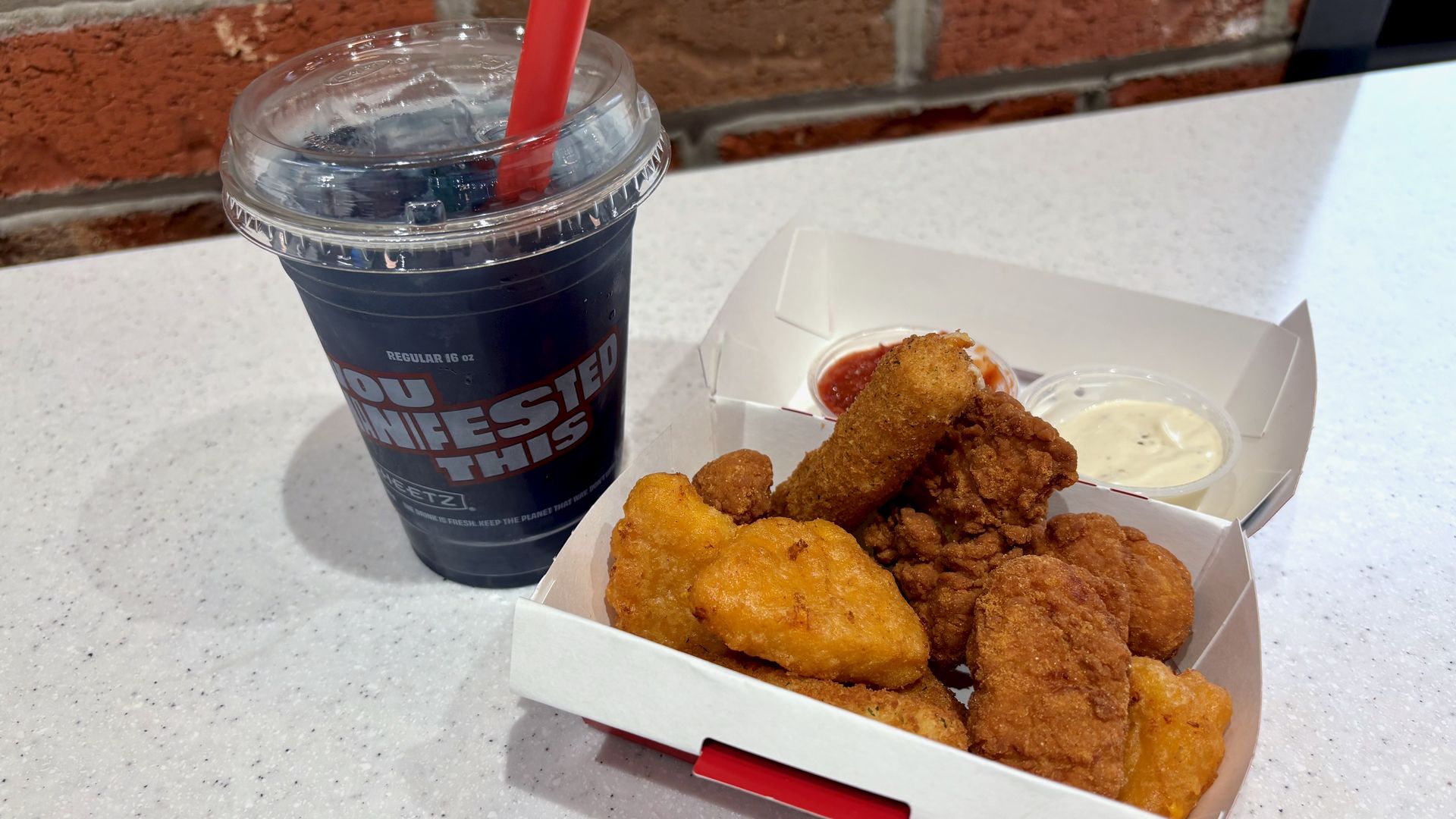 An app sampler in a paper box with mozzarella sticks, mac and cheese bites and boneless wings and a purple-colored berry lemonade