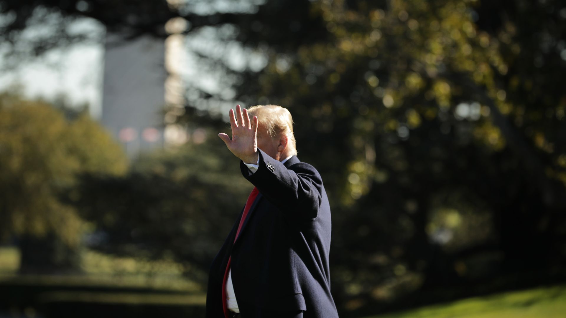 In this image, Trump waves at the camera while walking, which blocks out his face. The Washington Monument can be seen behind him.