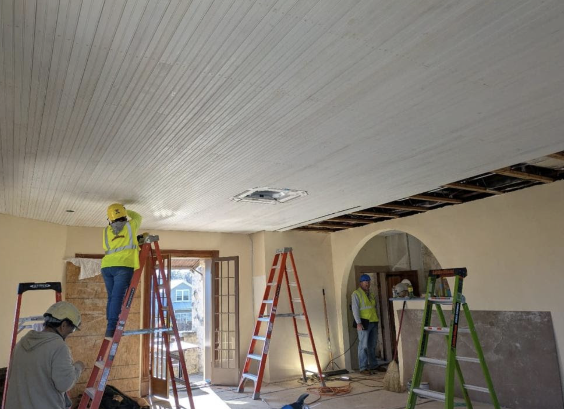Construction workers in safety gear renovating a room with beige walls, multiple ladders, and a partially exposed ceiling near an open door showing a blue house outside.