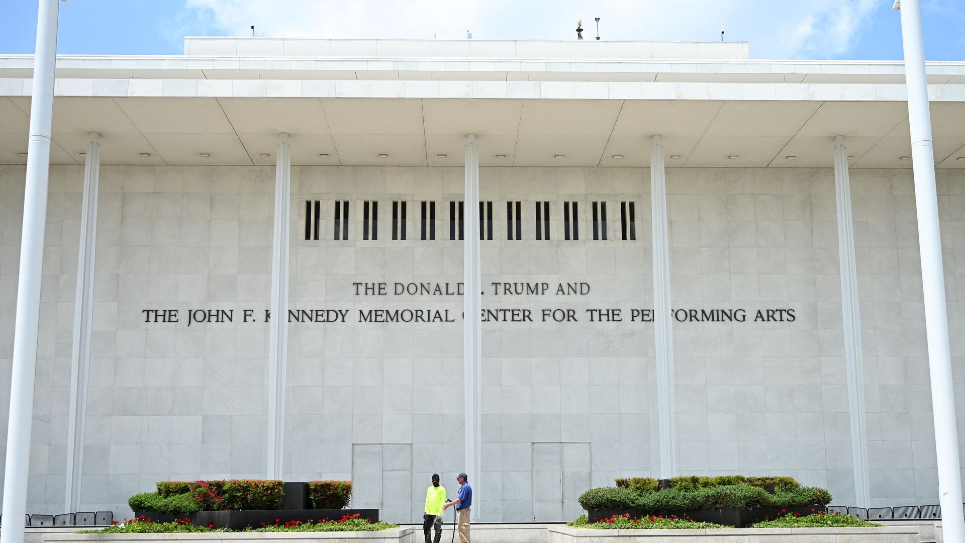 White stone building with tall columns and a wide overhang; inscription reads, "THE DONALD TRUMP AND THE JOHN F. KENNEDY MEMORIAL CENTER FOR THE PERFORMING ARTS." Two people stand by flower planters beneath a blue sky.
