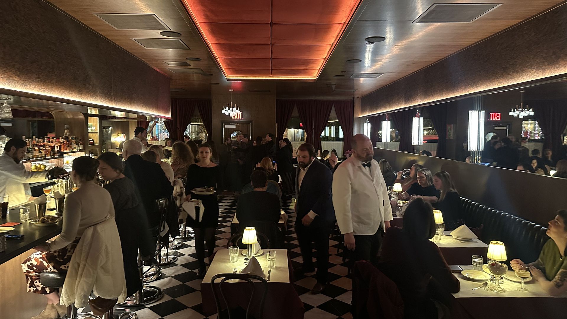 Busy upscale dining room with a long bar on the left, checkered floor, red-lit ceiling panels, and diners at tables along the right; waitstaff in white shirts and bowties moving about.