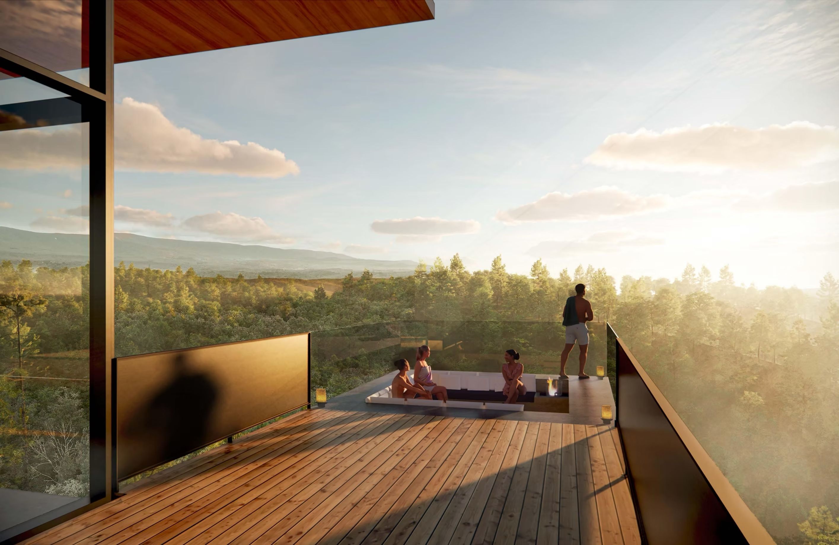 Modern glass balcony with a wooden deck overlooking a forest and distant hills at sunset. A white sectional sofa with people seated; one person stands at the railing.