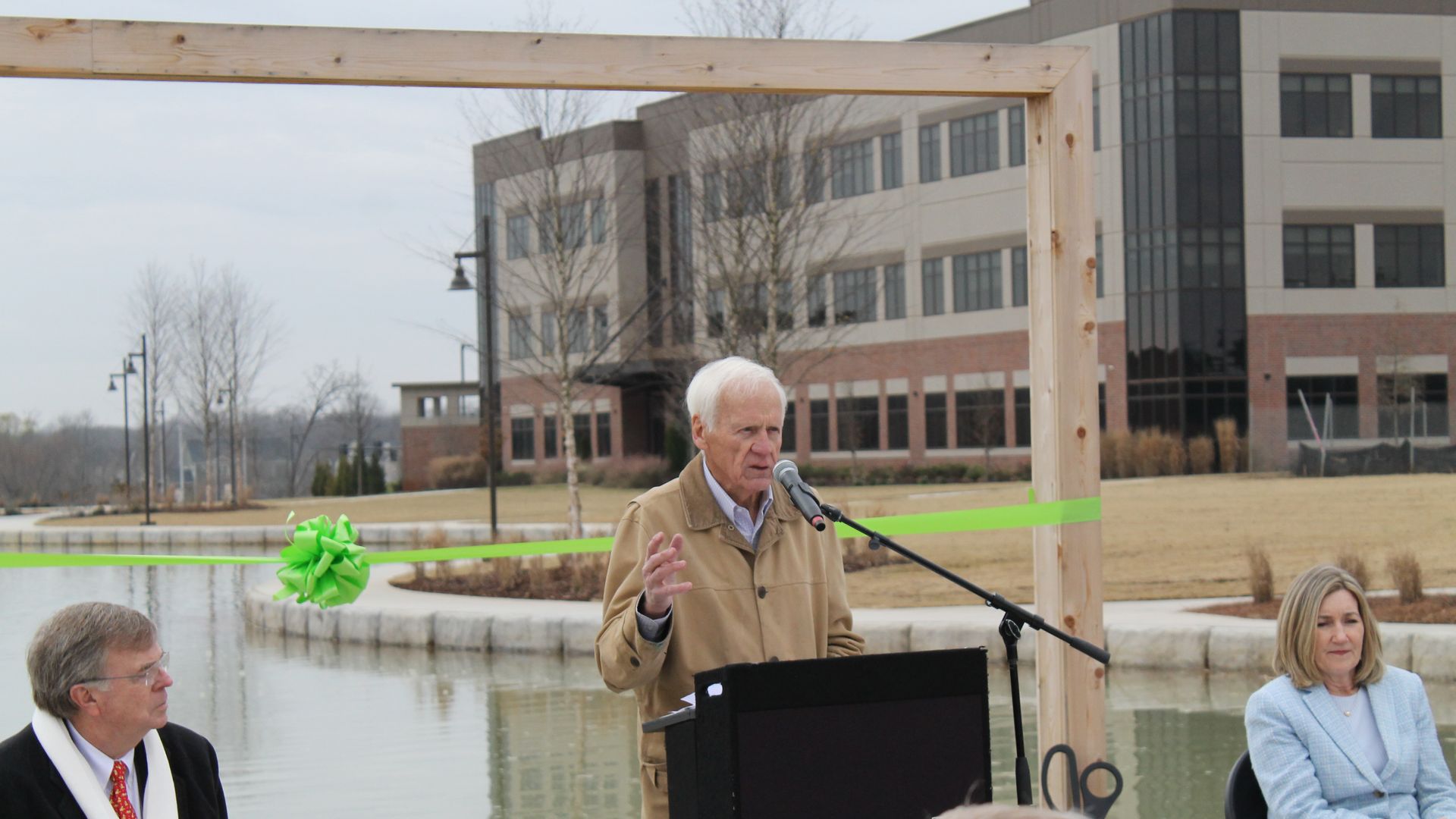 An older man in a beige jacket speaking at a podium outdoors near water, with a green ribbon and bow behind him, flanked by a man in a black coat with a white scarf and a woman in a blue blazer.