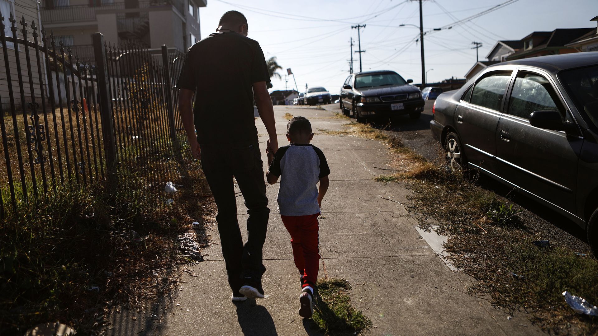A Honduran immigrant man and his young son hold hands with their backs to the camera walking down a sidewalk with a fence on one side.