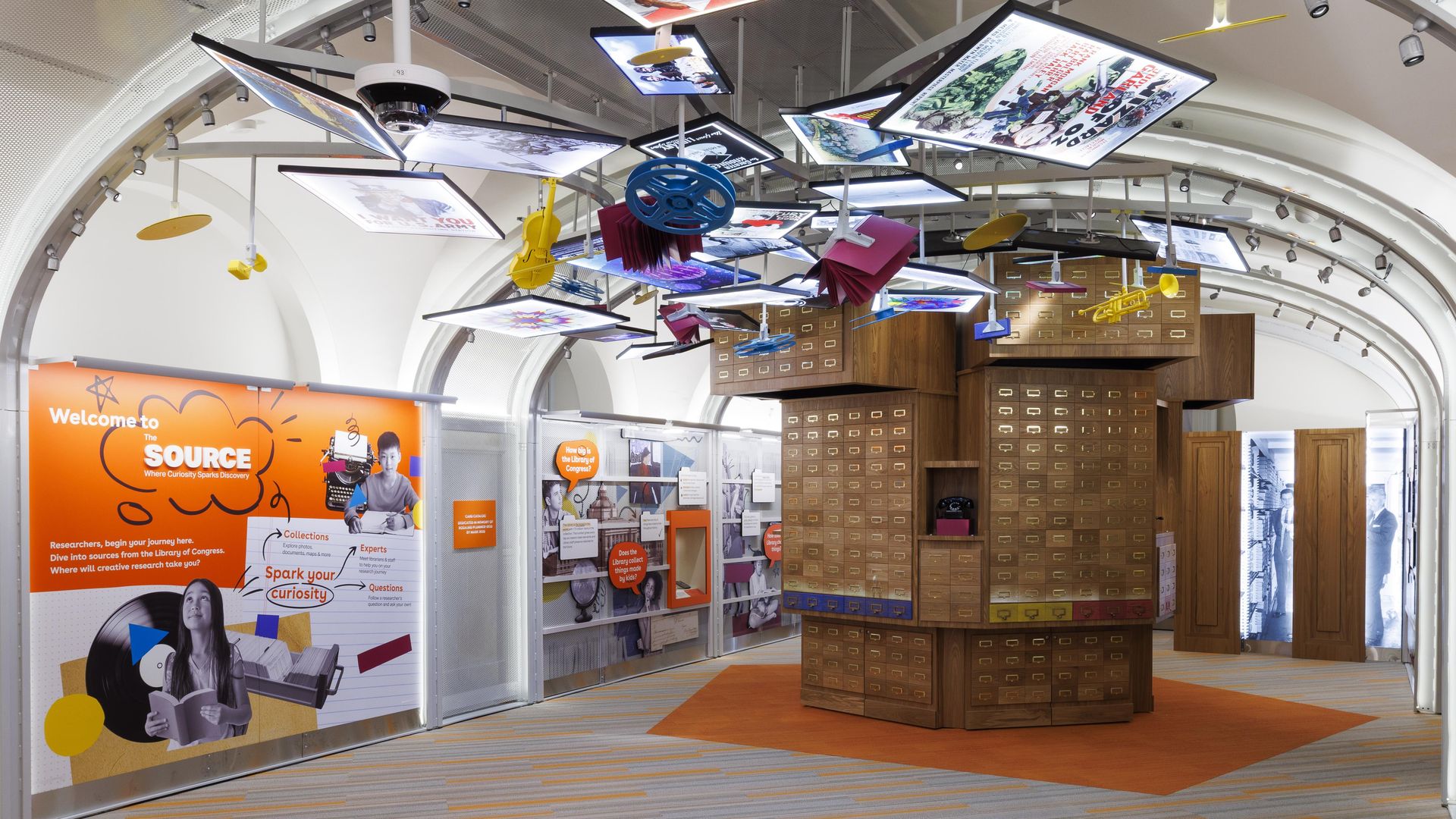 Bright exhibit space with many suspended screens displaying posters, a curved ceiling, and a tall stack of wooden filing cabinets; an orange panel reads "Welcome to The Source" with student imagery.