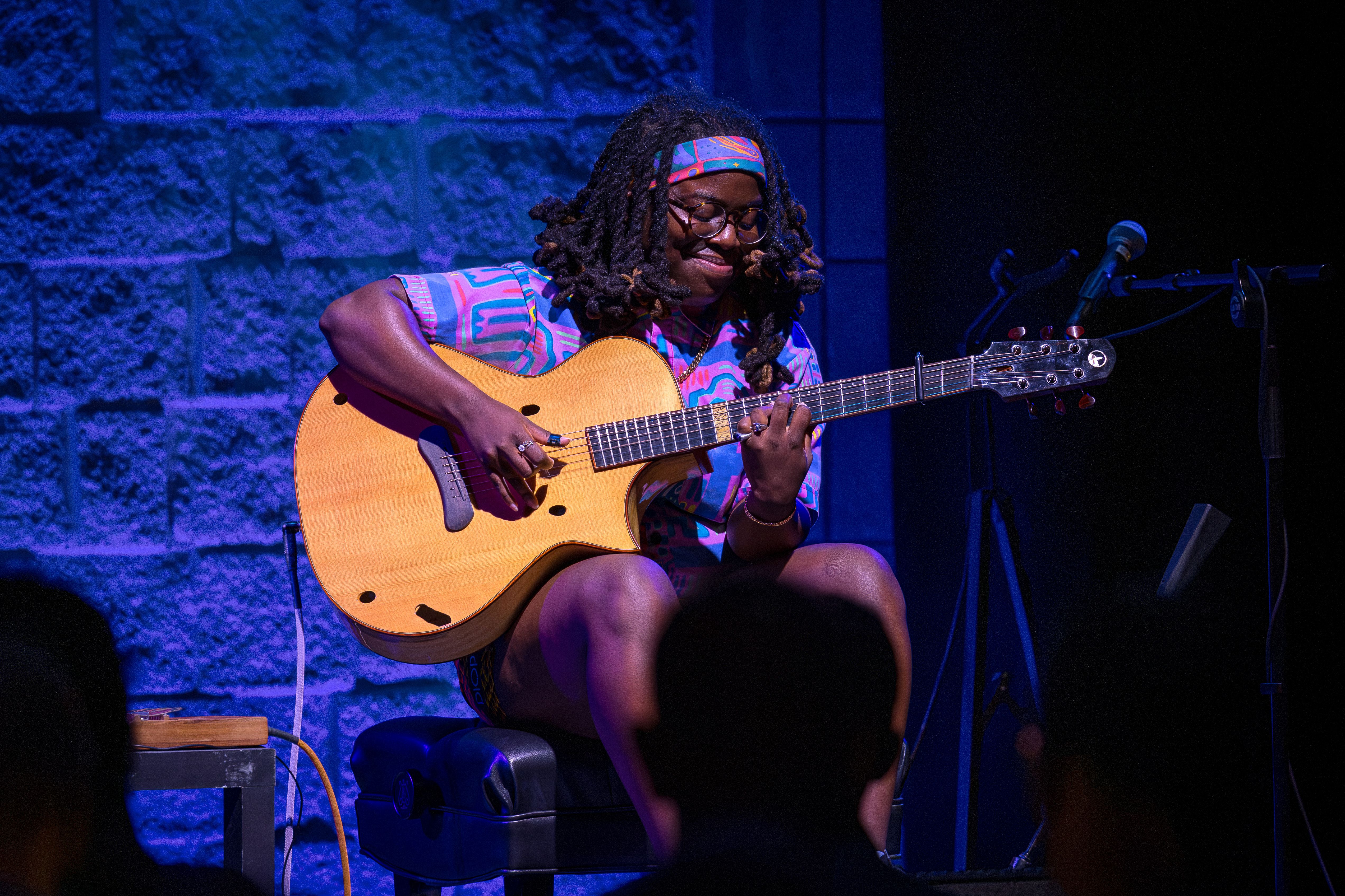 A musician with dreadlocks wearing a vibrant patterned shirt and headband, glasses, sits and plays an acoustic guitar on a blue-lit stage, with a microphone and stage gear nearby.