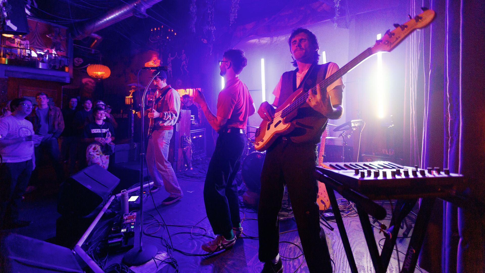 A band performs on stage with purple and blue lighting; one plays bass guitar, another guitar, and a third holds a microphone. Audience watches in a dimly lit venue with hanging decorations.
