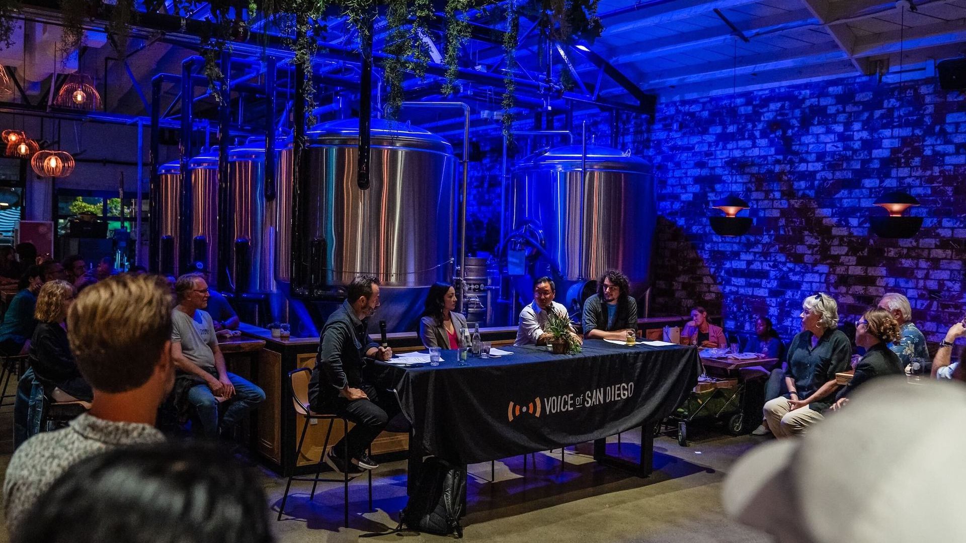 A panel of speakers sit at a table in front of crowd for an event at a brewery.