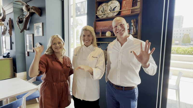 Two women and a man wave inside a restaurant.