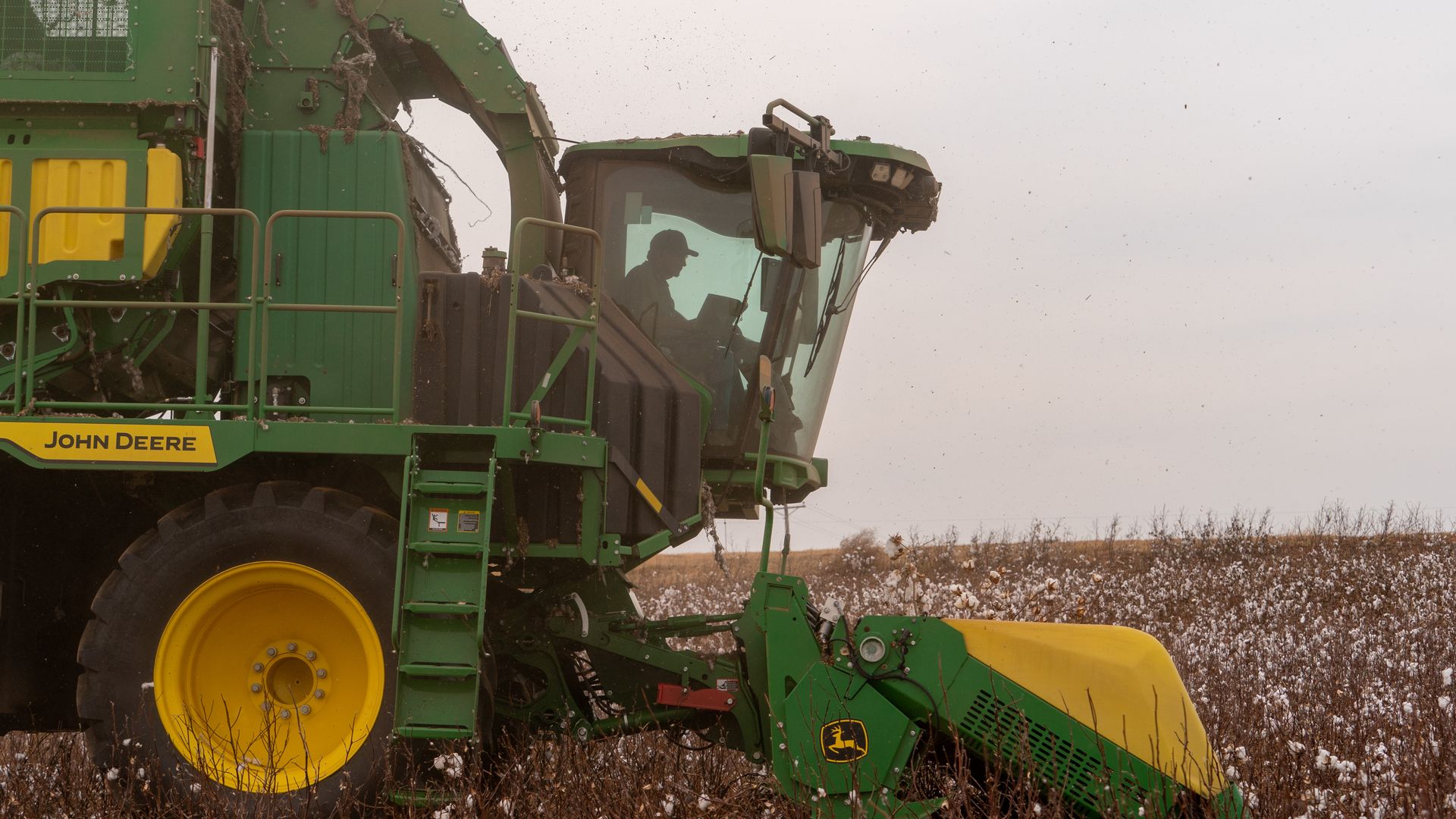 Green and yellow John Deere cotton harvester in a brown cotton field with white cotton bolls, a person visible in the cab, harvesting under a cloudy sky.