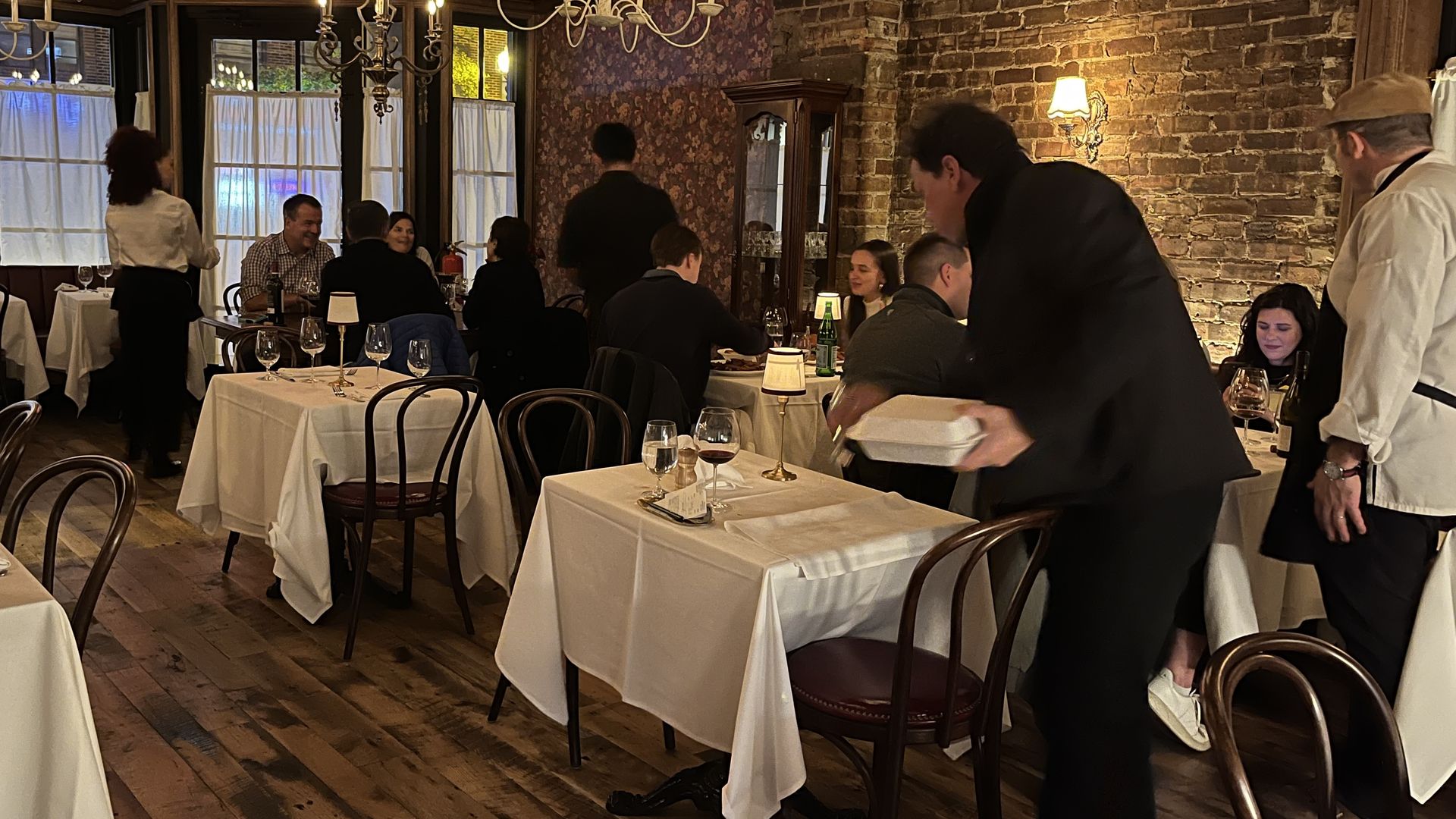 A warmly lit restaurant with brick walls and chandeliers. White-tablecloth tables are occupied by groups of diners; waiters move between tables on a wooden floor, with glasses and bottles visible.