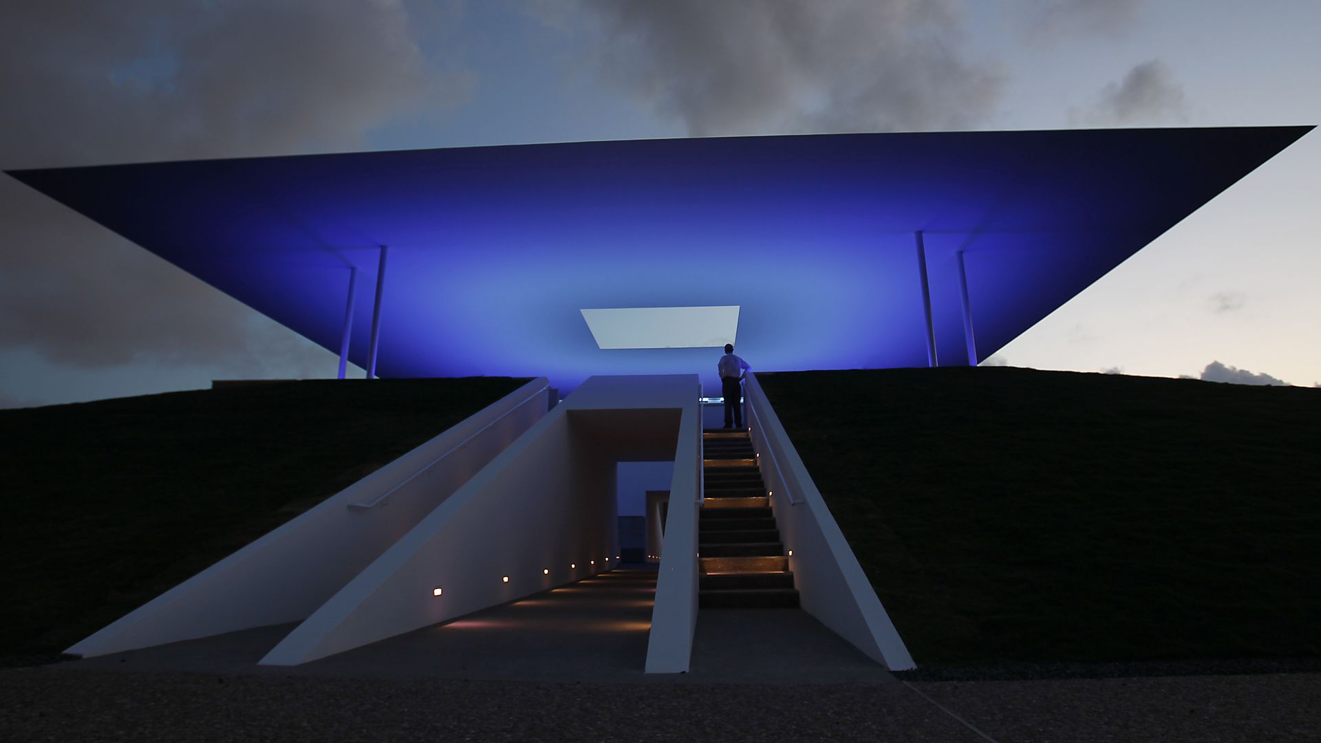 Silhouetted person on a white staircase leads to a large, blue-lit angular structure; a twilight sky with dark clouds forms the backdrop.