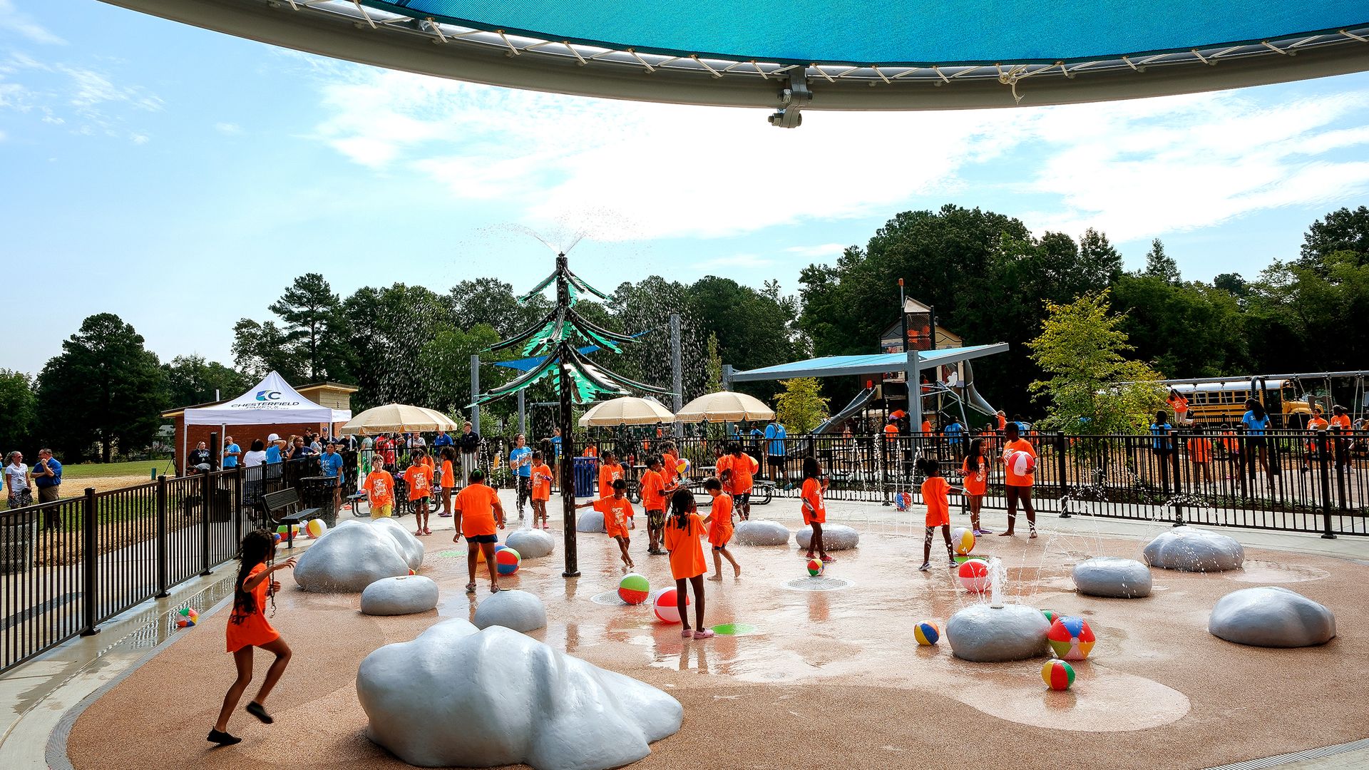 Children in orange shirts play on a splash pad with water jets, gray rock-like sculptures, and colorful balls under a large blue shade sail; a fence, tents, trees, and a playground are in the background.