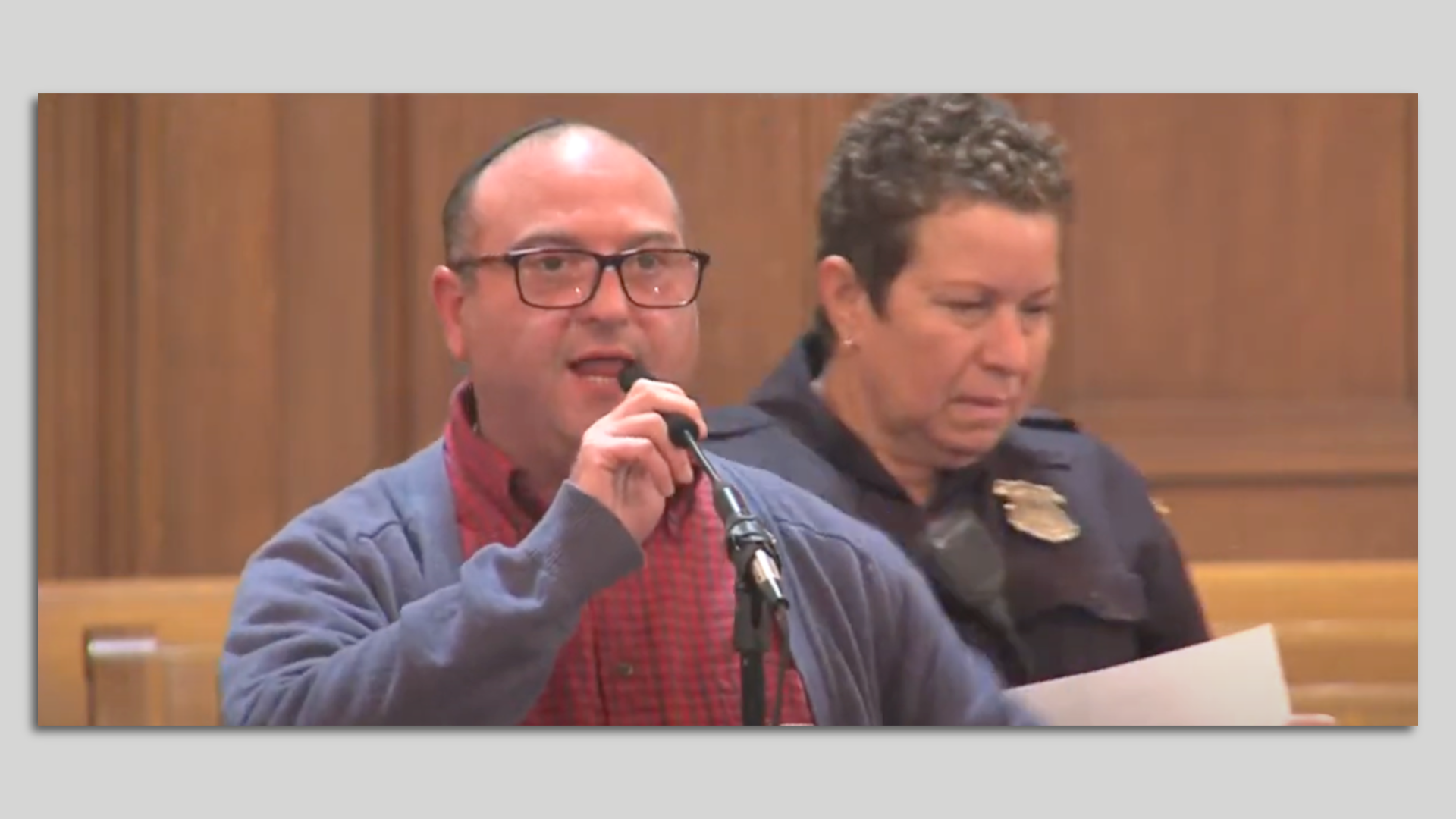 A man in glasses holding a microphone in a wood-paneled legislative chamber with a police officer immediately behind him.