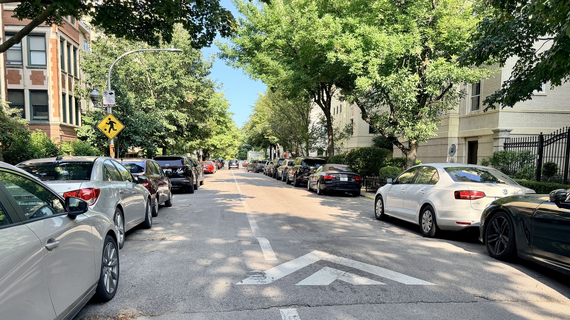 Sunlit street with parked cars on both sides, green trees overhead creating a leafy canopy. A yellow pedestrian crossing sign sits near the curb as brick and beige buildings frame the block.
