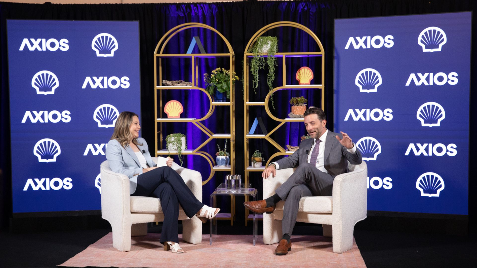 Two people sit in white armchairs on a stage with blue Axios banners and Shell logos; a gold decorative shelf with plants stands between them as they chat on a pink rug.