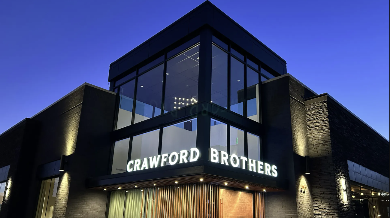 Modern building corner at dusk with large glass windows and illuminated sign reading "CRAWFORD BROTHERS" against a clear deep blue sky.