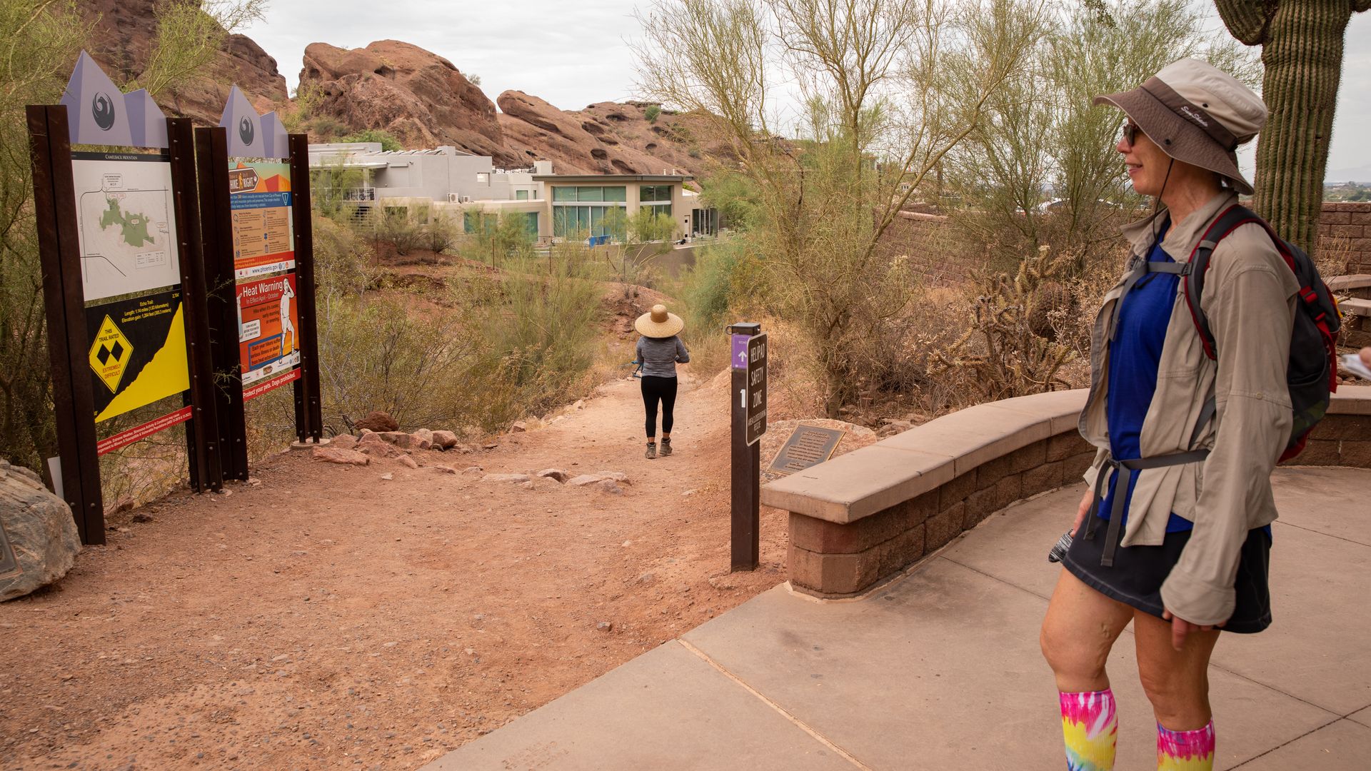 Two hikers in desert terrain near trail signs; one wears a wide-brimmed hat walking on dirt path, the other in a sun hat, blue shirt, and colorful tie-dye socks stands on paved area with cactus nearby.