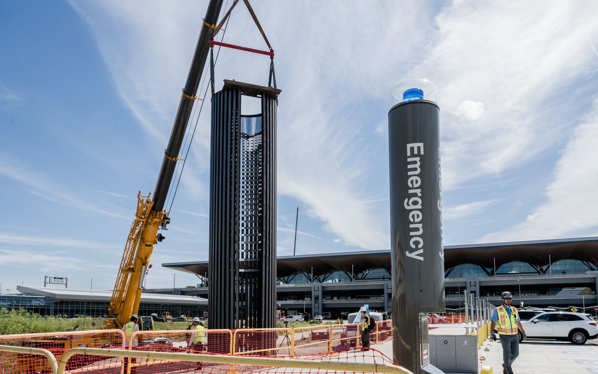 Construction site with a large black ribbed cylinder hoisted by a yellow crane. A blue beacon atop a gray emergency pillar stands to the right; orange fencing and workers in hi-vis vests nearby.