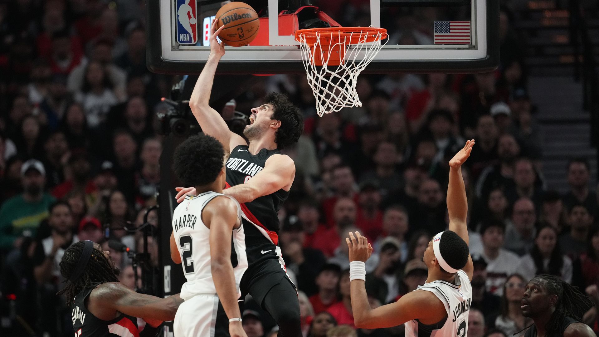 Portland player in a black jersey dunks near the rim, gripping an orange ball; defenders in white reach up as the crowd watches, NBA logo on the backboard.